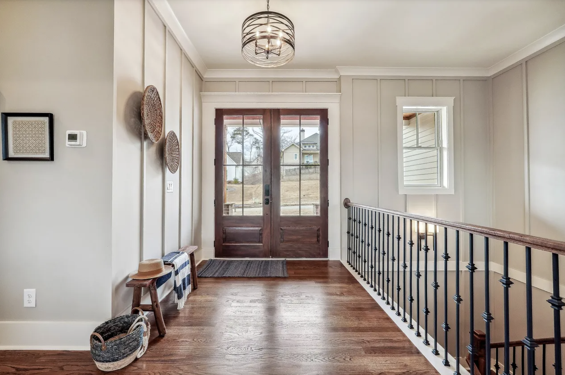 Entryway with wooden front door, window, and staircase with black railing, decorated with woven baskets and framed art on a beige wall, and a small bench with a striped cloth and a straw hat.