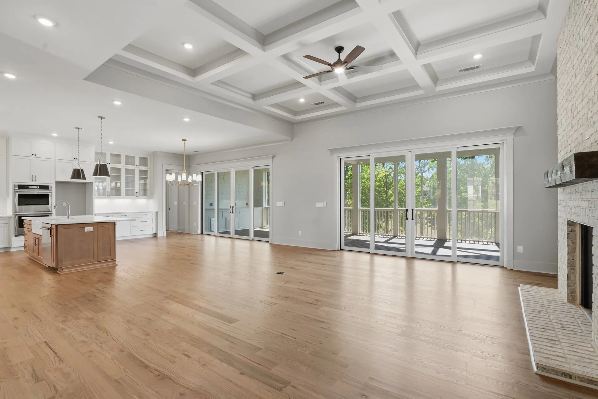 Empty living room with wooden floors, white walls, large sliding glass doors leading to a balcony, kitchen in the background with white cabinets, island with a sink, and hanging pendant lights, ceiling with recessed lights and ceiling fan.