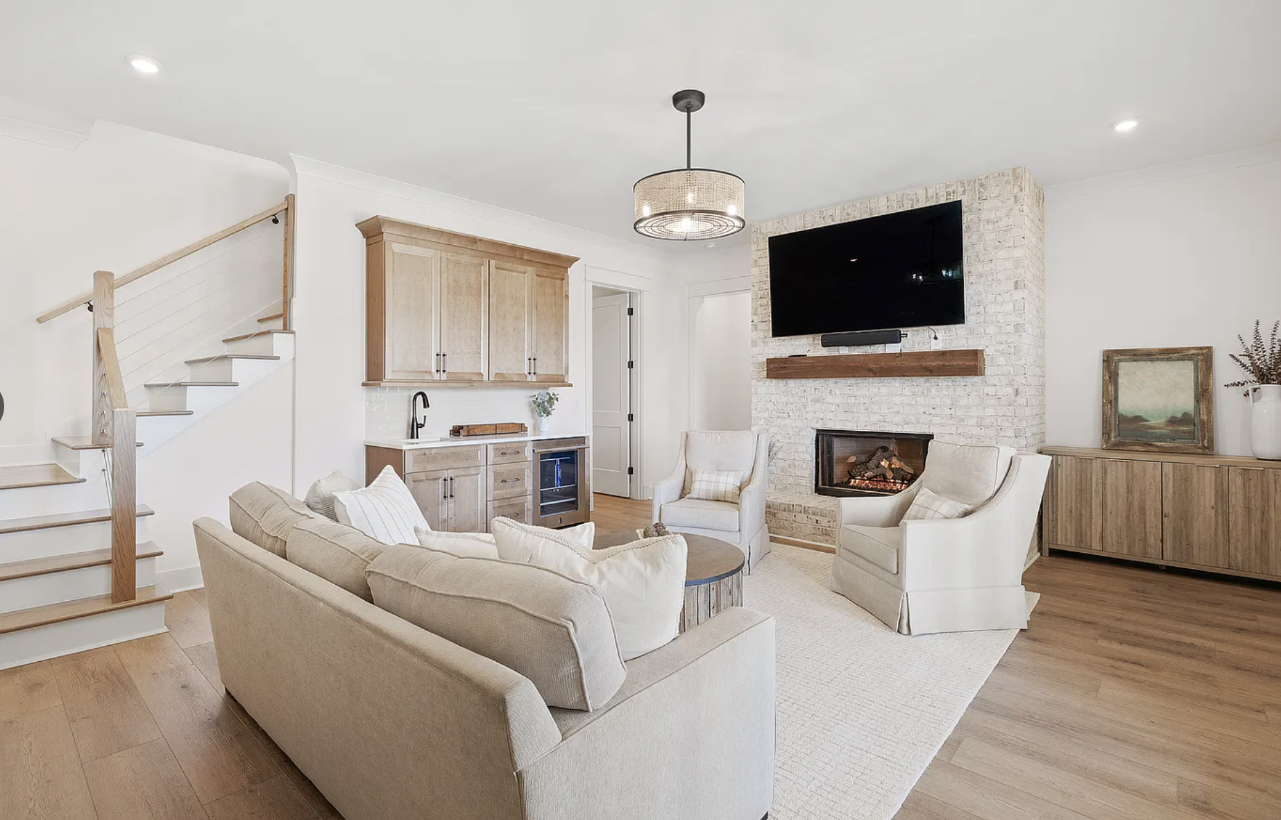 Living room with white brick fireplace, wall-mounted TV, beige couch, two white armchairs, wooden cabinet, and staircase on the left.