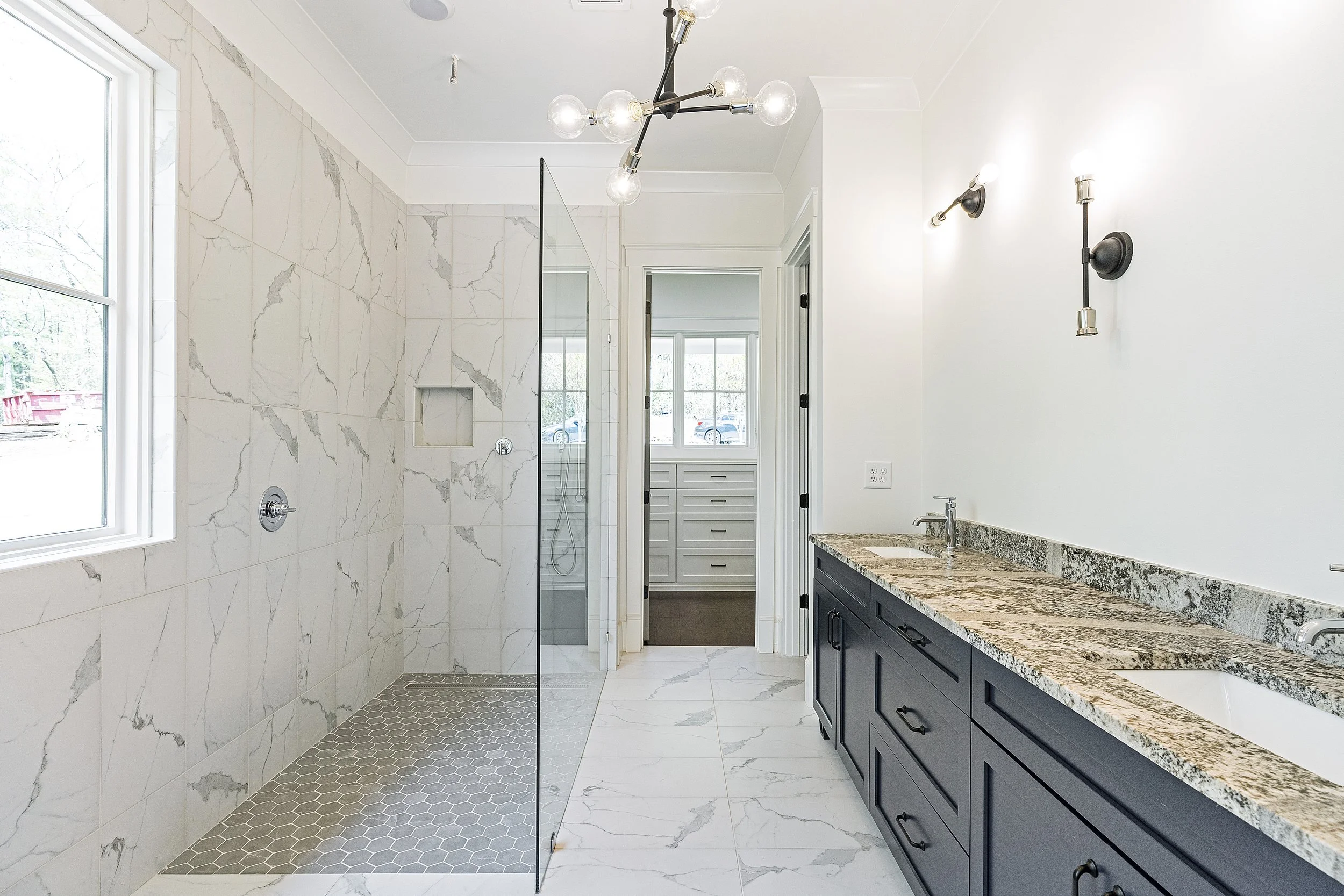 Bathroom with a marble shower, gray hexagonal floor tiles, a large window, a modern chandelier, and a gray vanity with a stone countertop.
