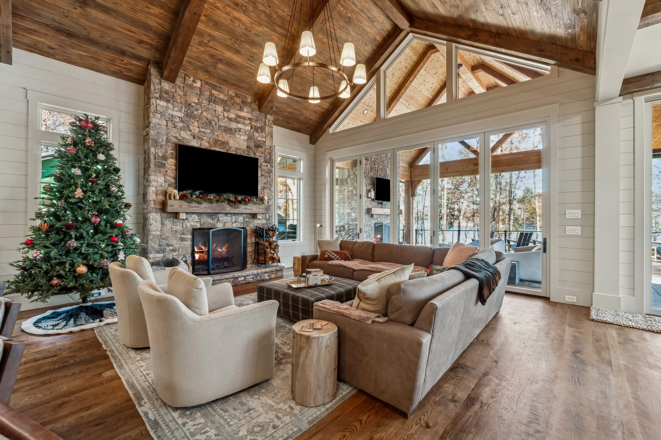 Interior of a living room with a Christmas tree, a stone fireplace with a TV on it, large windows, and wooden vaulted ceilings.