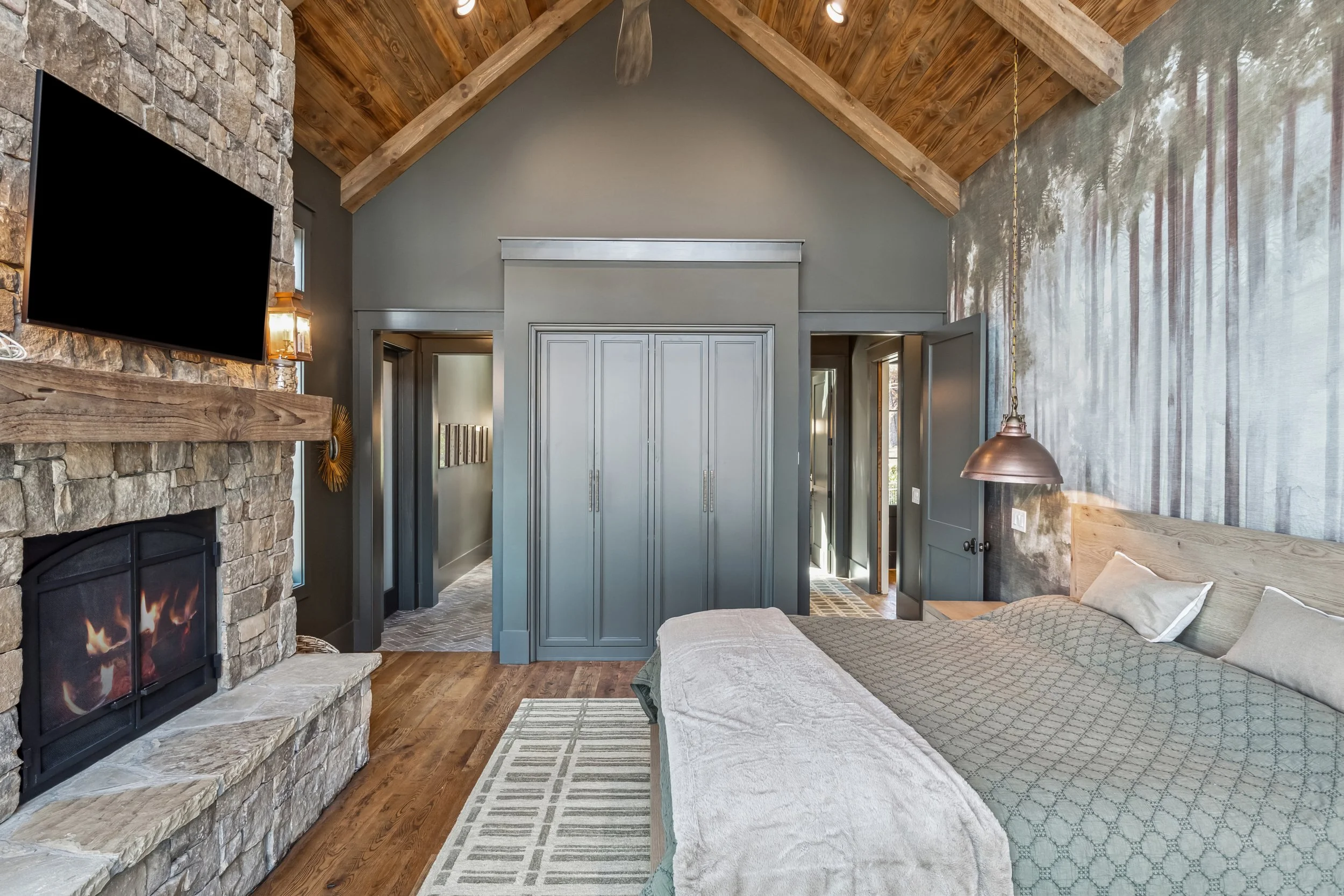 A cozy bedroom with a stone fireplace on the left, a wooden vaulted ceiling, a large wall with a mural of trees, a bed with light-colored bedding, and a hanging copper pendant light on the right.