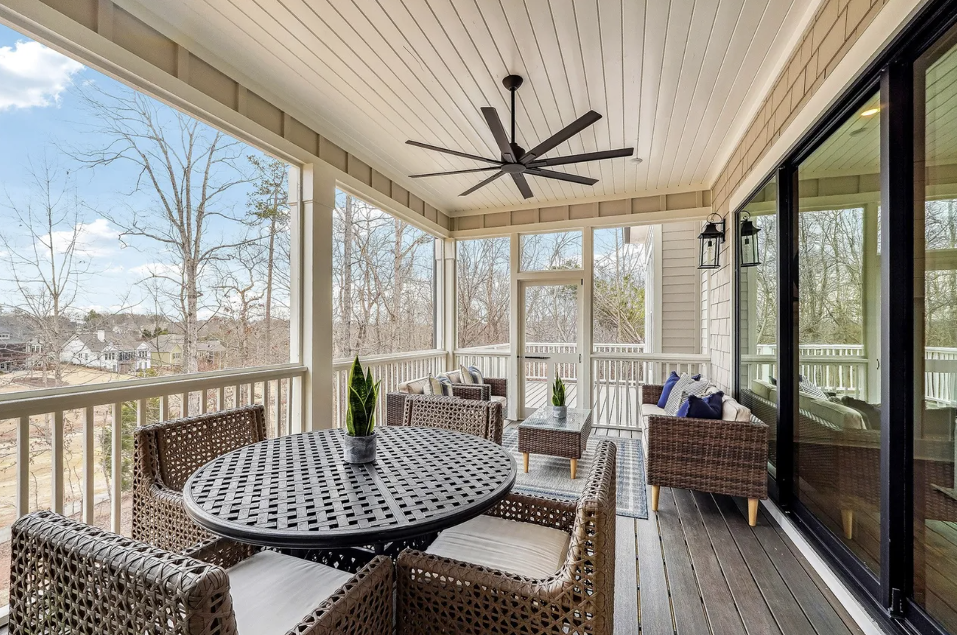 Enclosed porch with patio furniture, including a round table, chairs, and a sofa with cushions, overlooking a backyard with leafless trees and houses, with a ceiling fan and hanging lanterns.