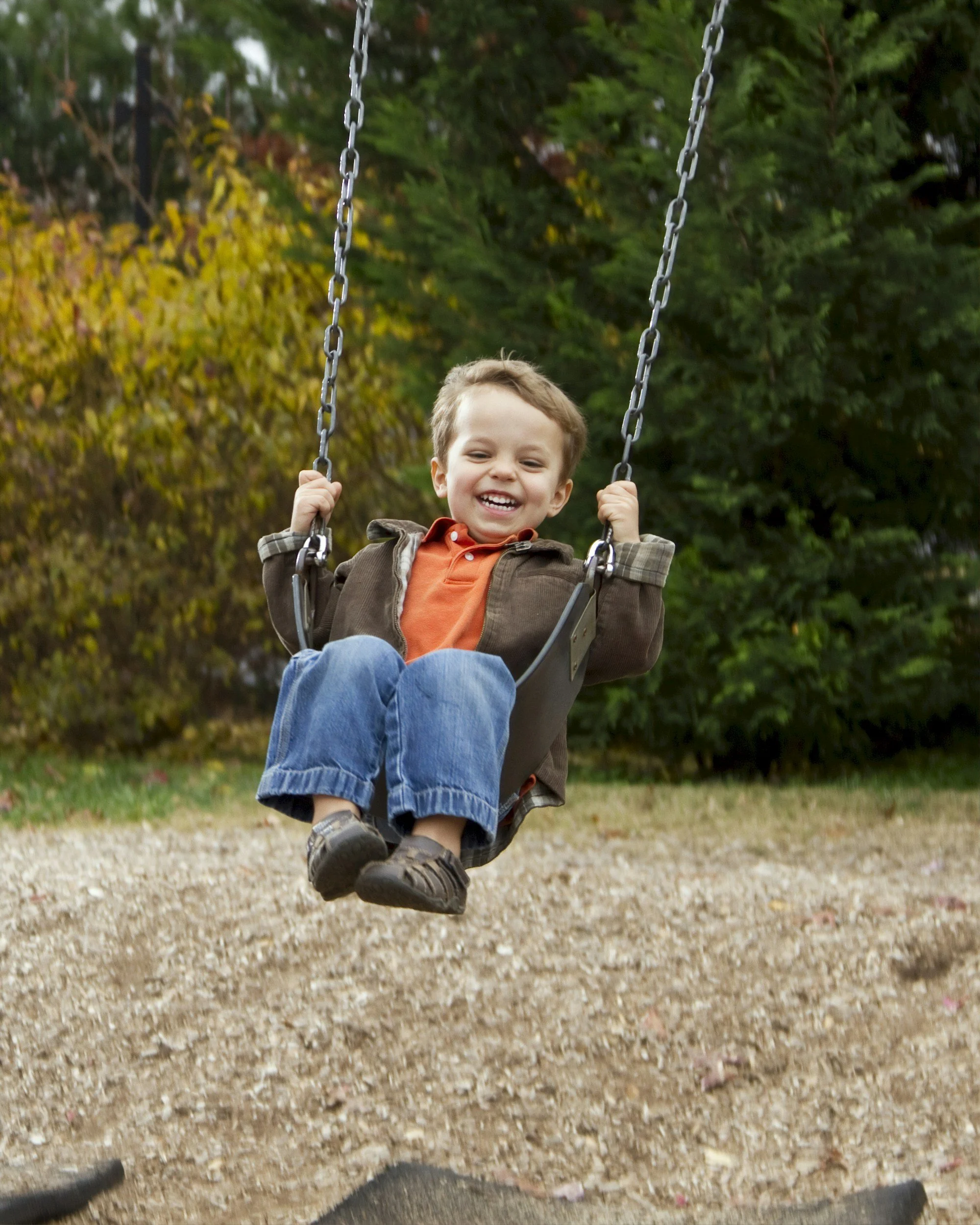Young boy with brown hair smiling as he swings on a playground swing set, surrounded by green and yellow trees.