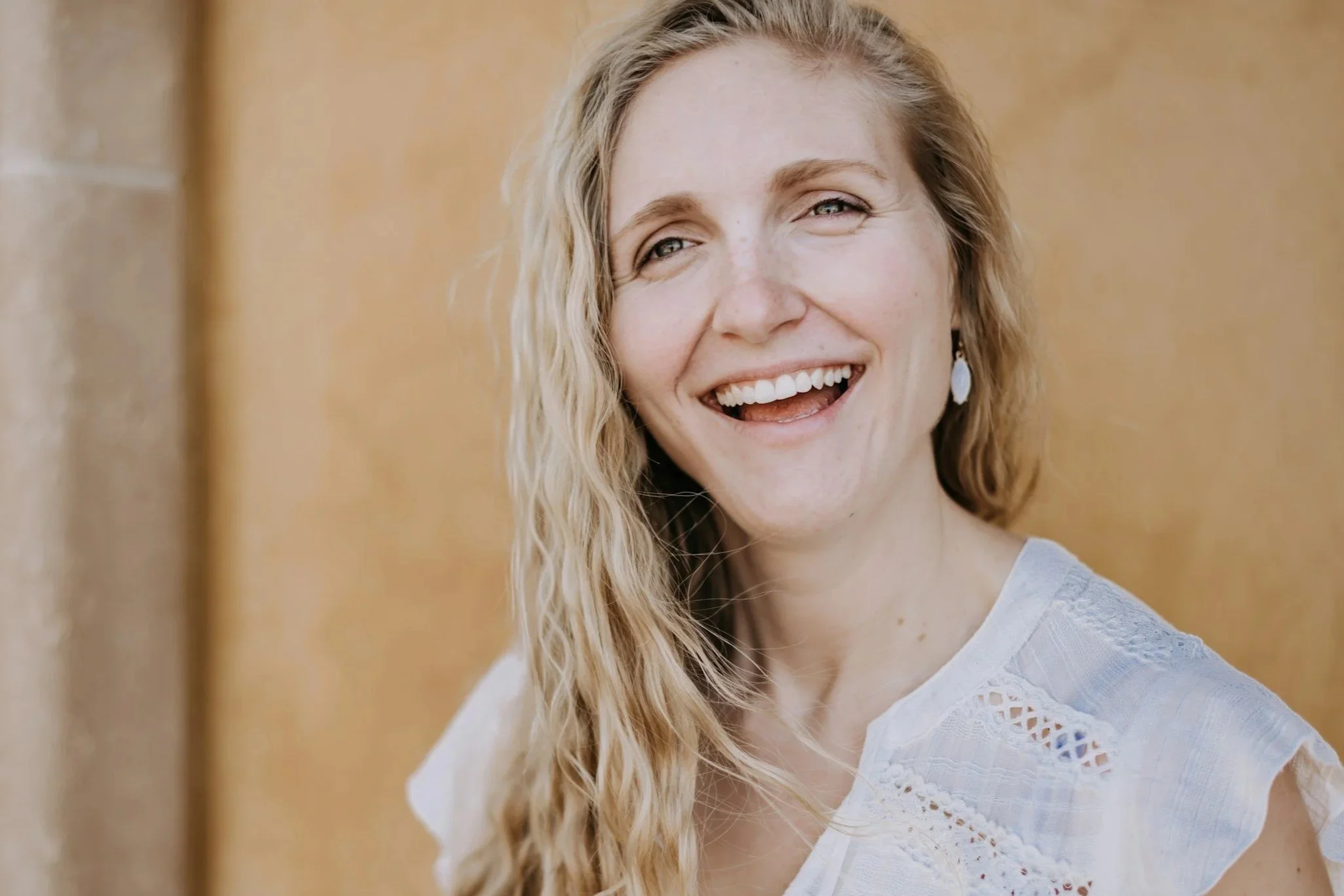 A woman with blonde, curly hair smiling with white teeth, wearing a white lace top, earrings, standing against a wooden background.