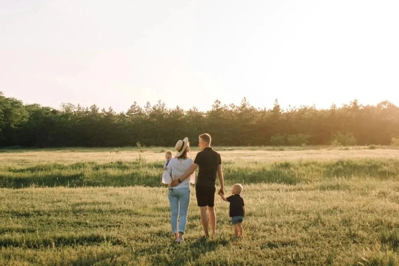 A family walking hand in hand across a grassy field at sunset, with a woman carrying a toddler and a young boy holding her hand.