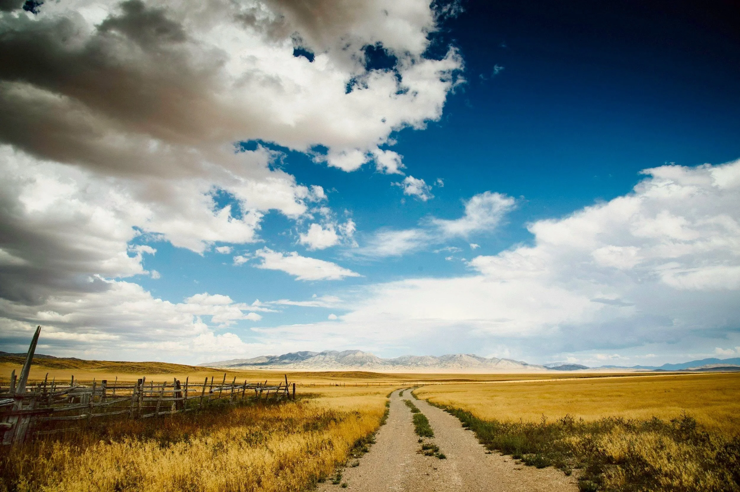 A dirt road winding through an open field with tall grass, a wooden fence on the left, and mountains in the background under a partly cloudy sky with patches of blue.