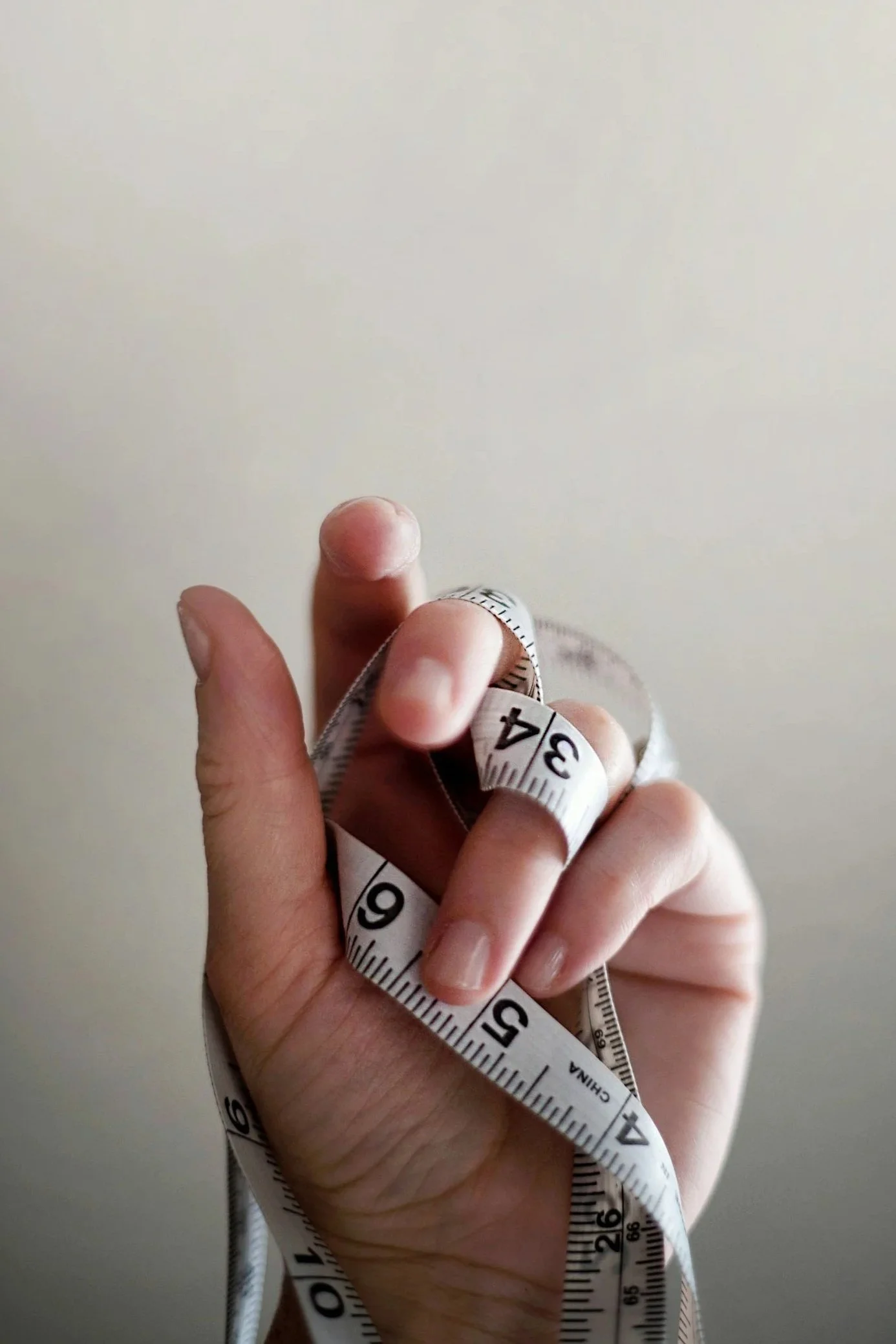 A person holding a white measuring tape with black numbers, with some fingers visible, against a plain light-colored wall.