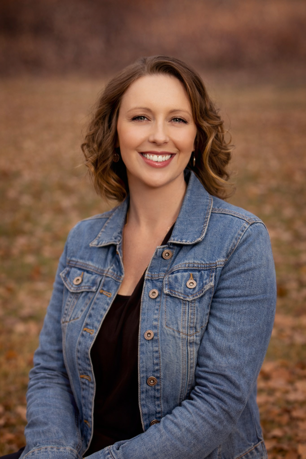 A smiling woman with shoulder-length wavy brown hair, wearing a denim jacket over a black top, outdoors during fall with fallen leaves on the ground and a blurred background.