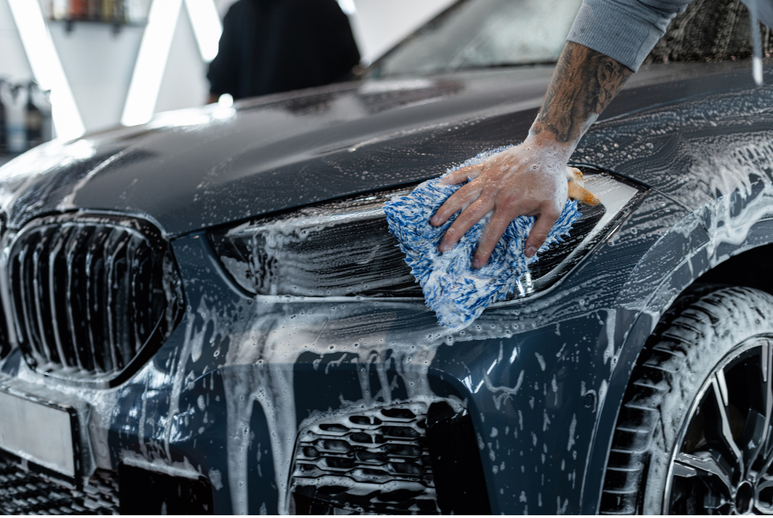 A person cleaning the front of a black car with a blue sponge and soapy water.
