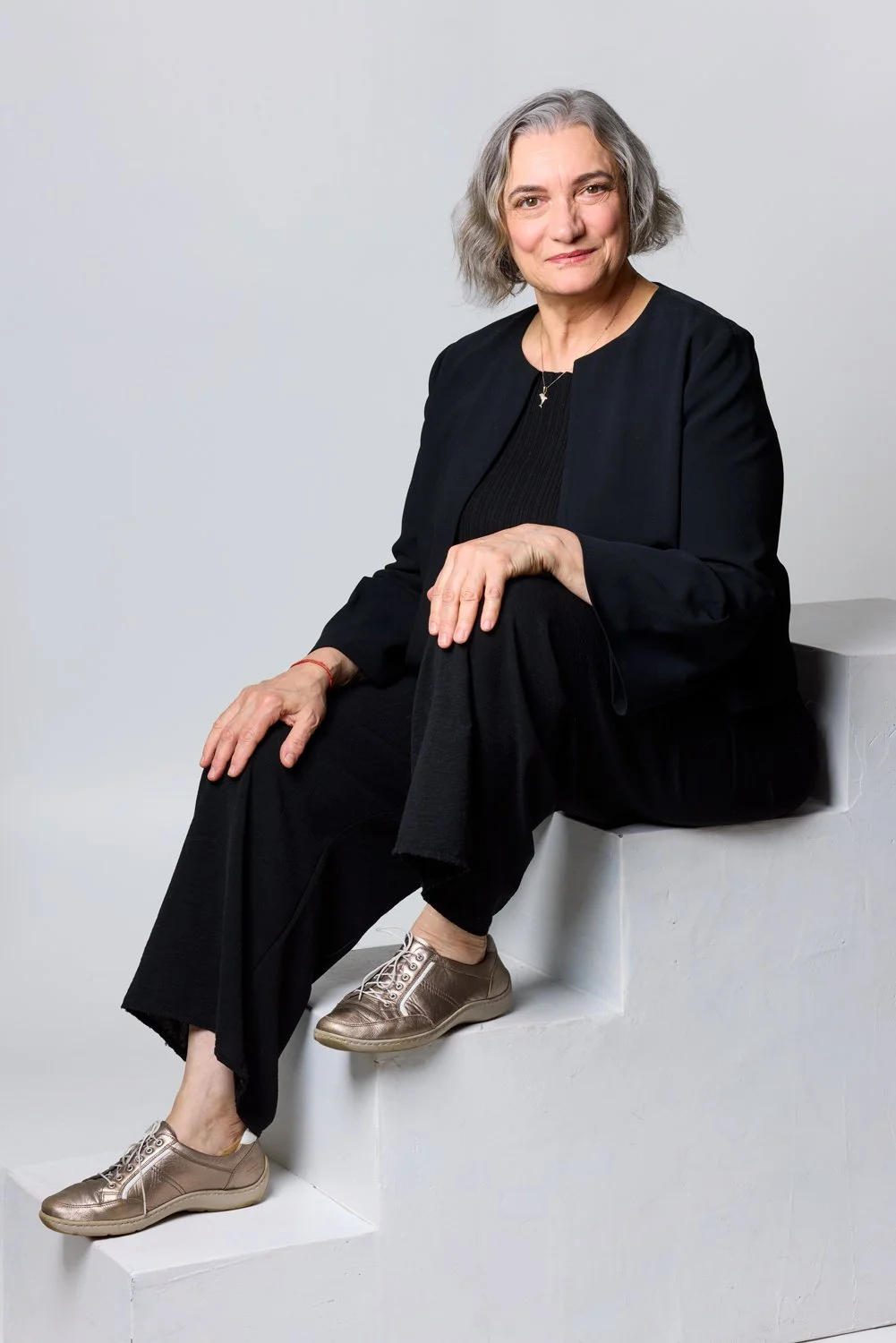 A mature woman with gray hair sitting on a white step against a plain light gray background, wearing a black outfit and gold sneakers, looking at the camera with a slight smile.