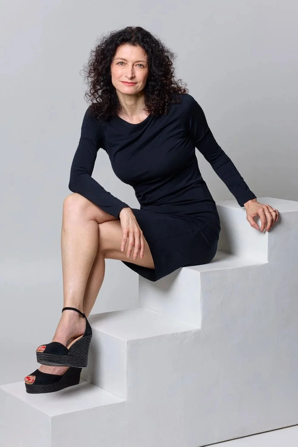 A woman with curly dark hair wearing a black dress and black wedge sandals sitting on white steps against a plain grey background.