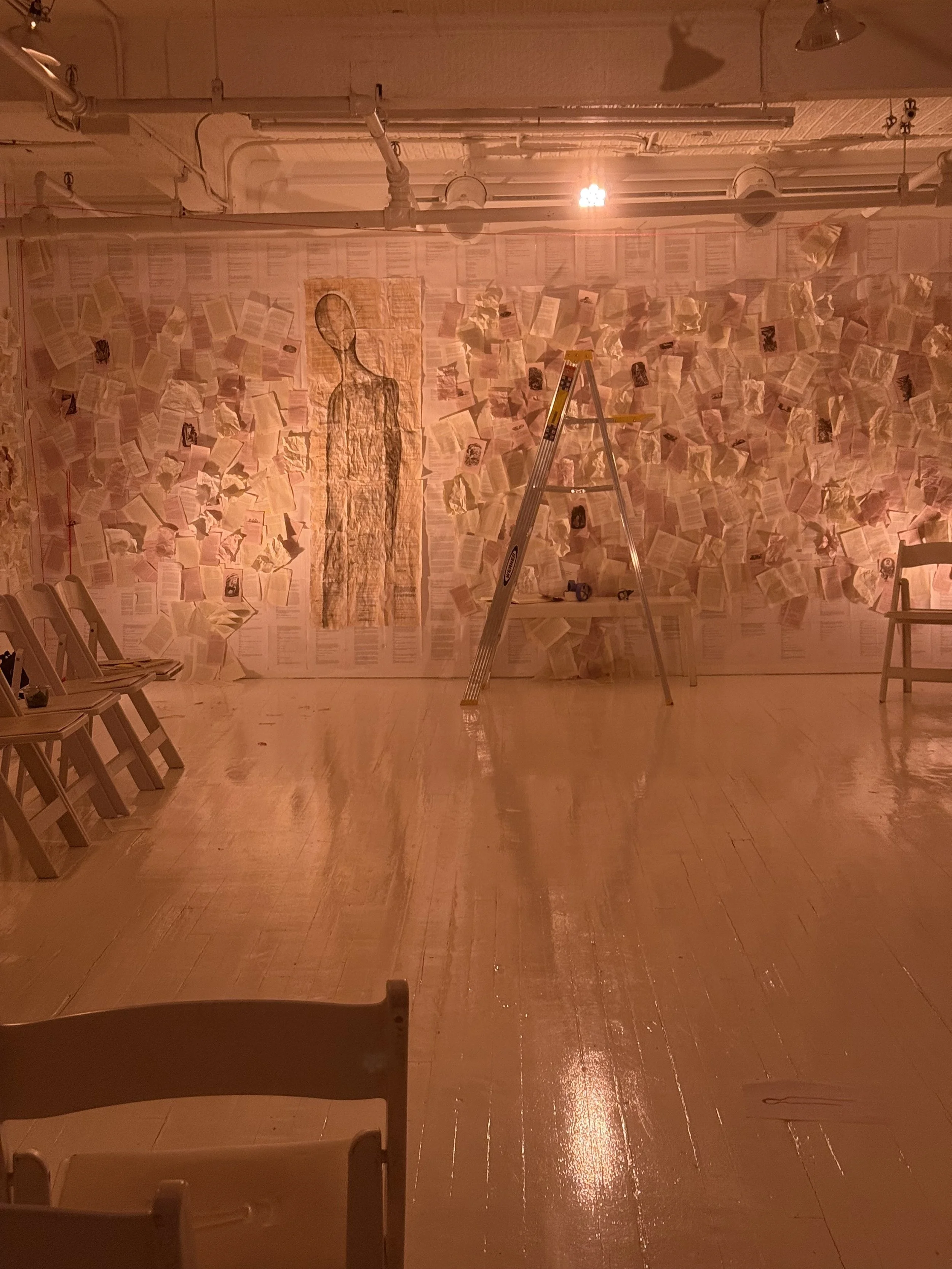 An art installation featuring a wall covered with scattered open books, with a large outline of a human figure hanging among them, illuminated by a soft warm light in a room with a wooden floor and chairs.