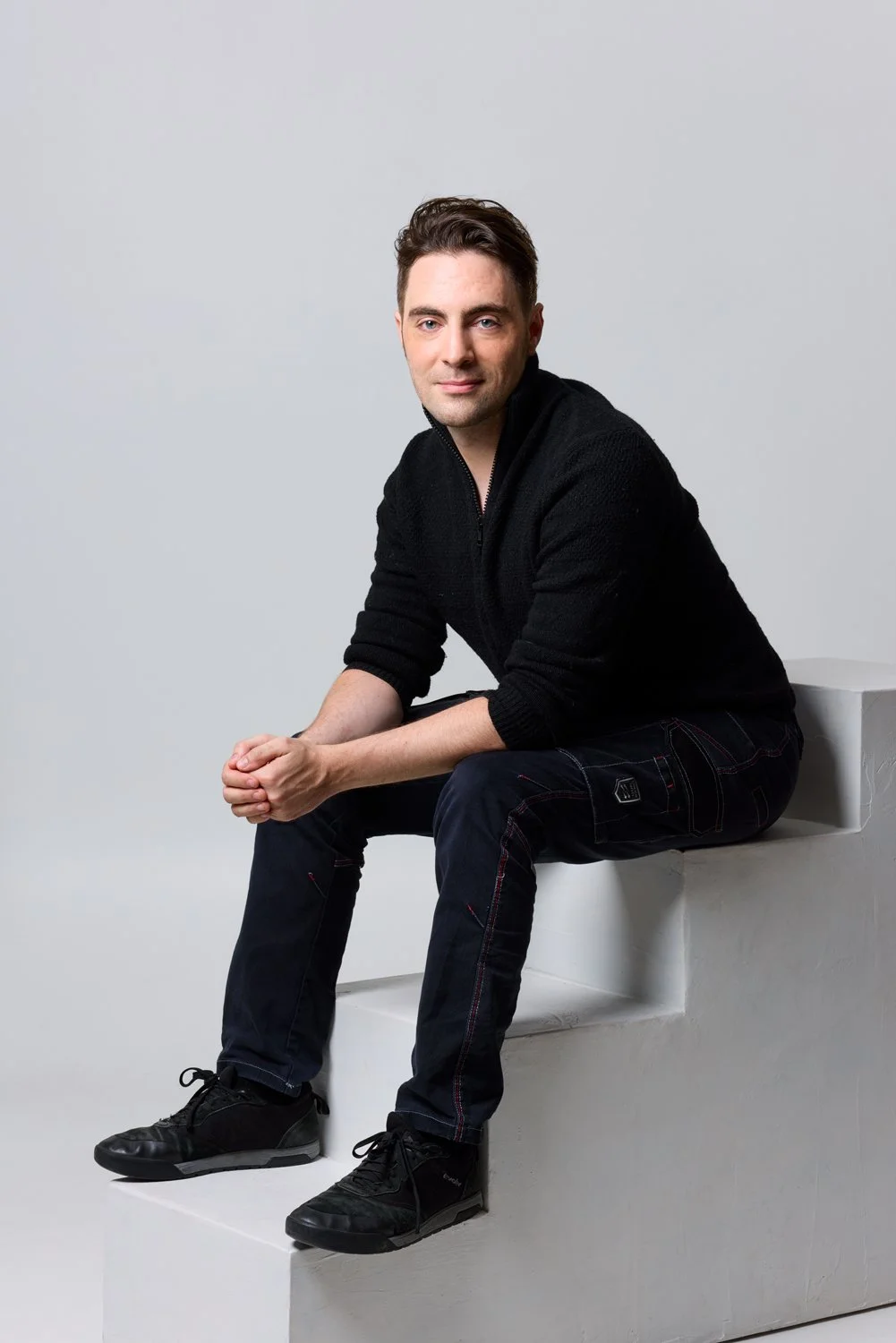 A young man sitting on a white step, wearing a black sweater, dark jeans, and black shoes, with a neutral expression and looking at the camera against a plain white background.