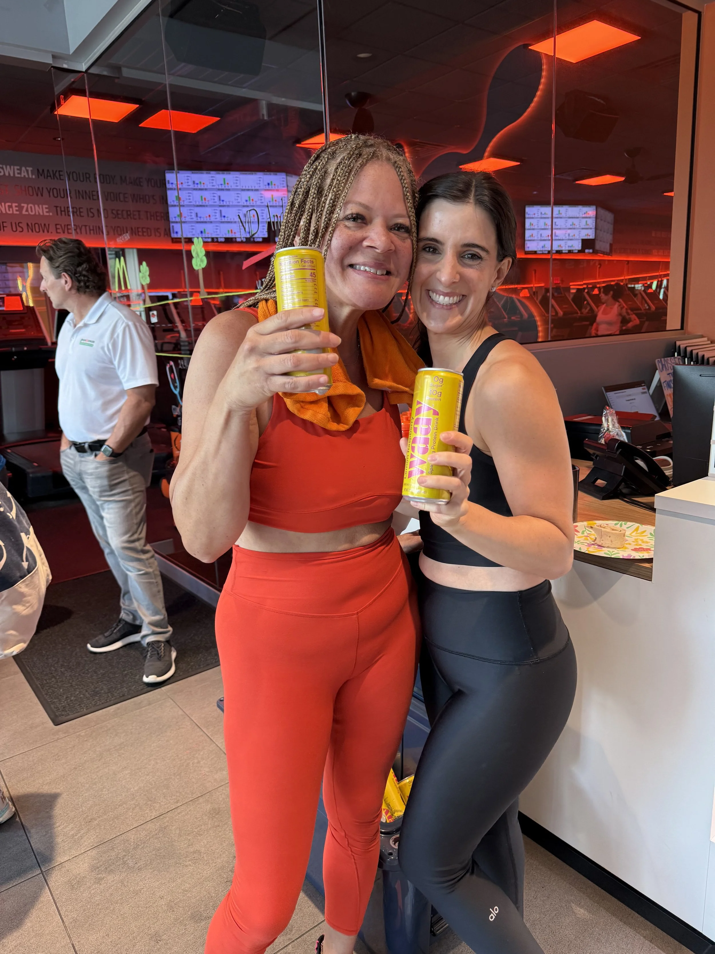 Two smiling women in fitness attire holding canned energy drinks in a gym or fitness studio with mirrors and workout equipment in the background.
