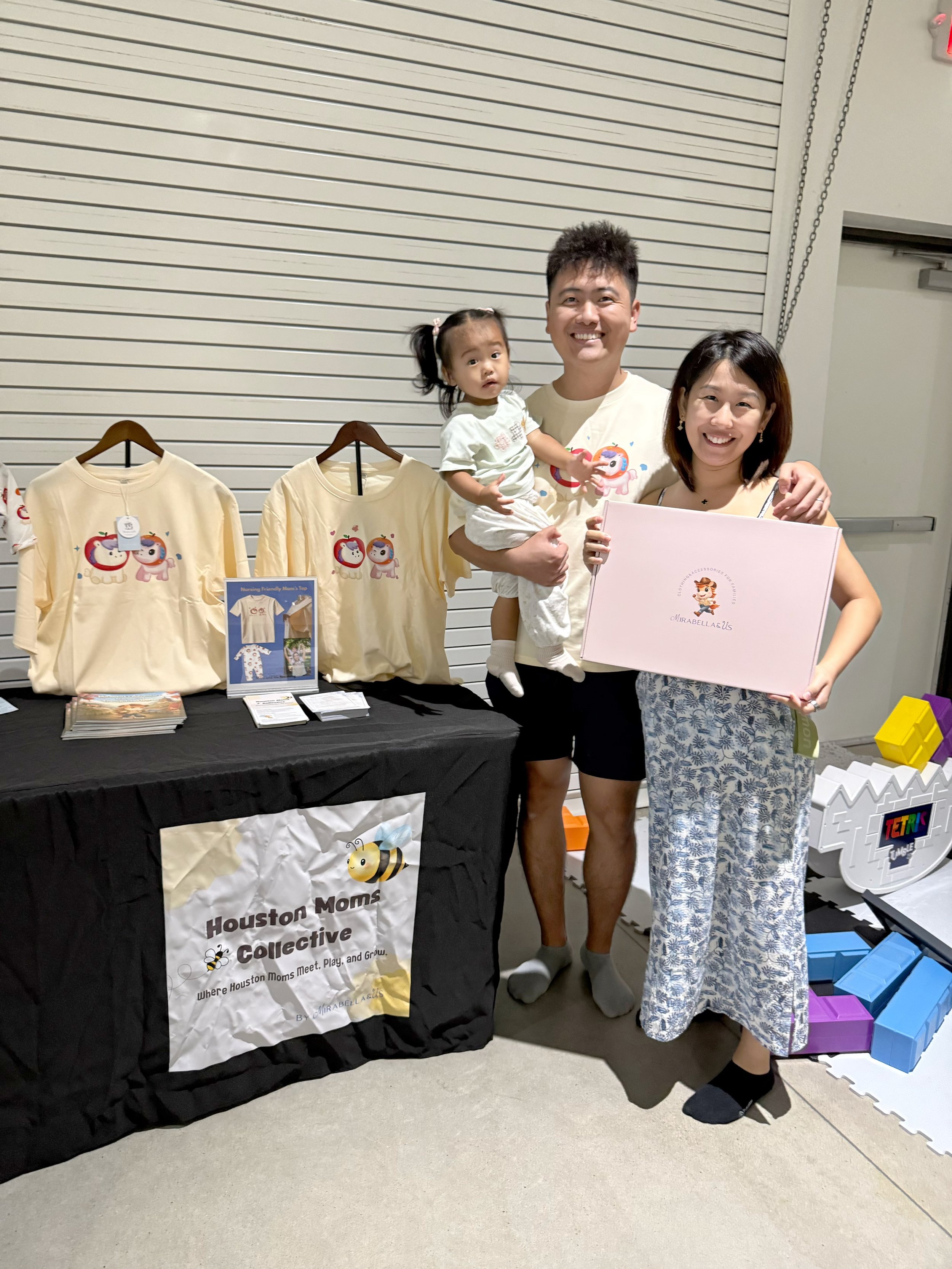 A family of three smiling and holding an envelope, standing behind a table with promotional materials for the Houston Moms Collectives in an indoor setting. The table displays yellow T-shirts with a cartoon lion and unicorn, and a banner with the gro