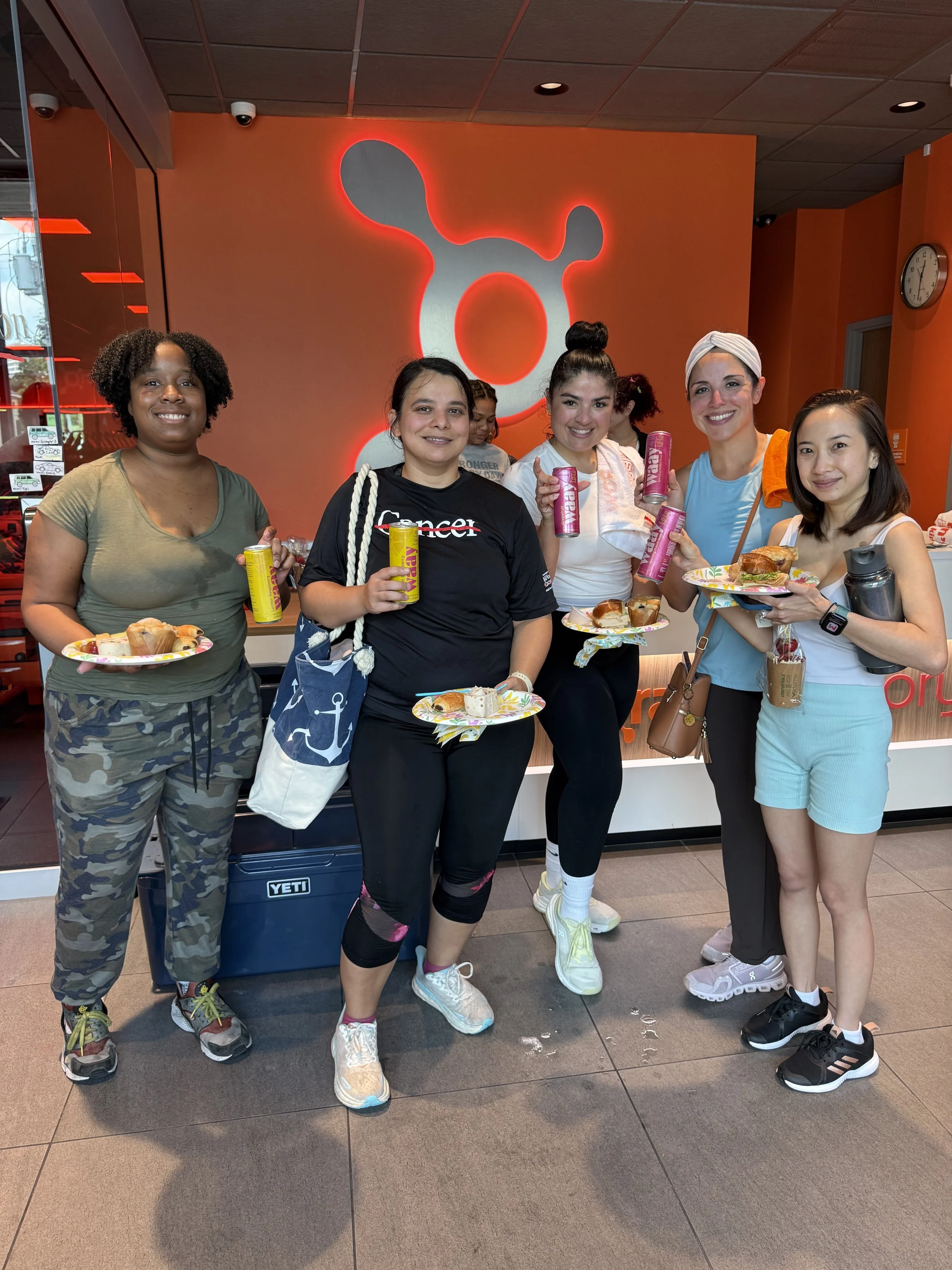 A group of five women standing together inside a building, holding plates of food and drinks, smiling at the camera. Behind them is an orange wall with a large illuminated graphic design.