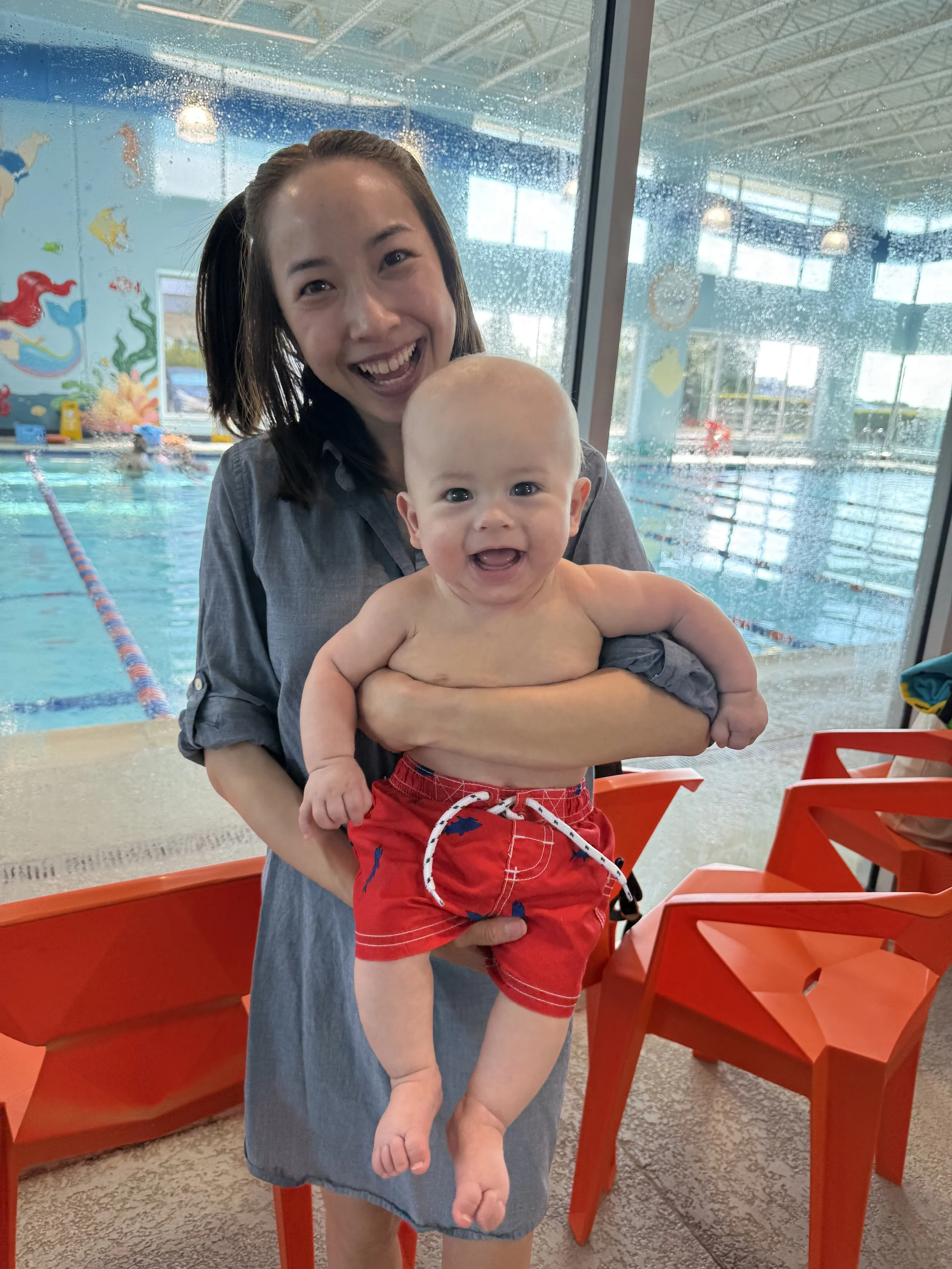 A woman smiling and holding a baby boy at an indoor swimming pool area. The woman has dark hair and is wearing a denim dress, while the baby is shirtless, wearing red swim shorts with a drawstring. The background features a swimming pool with water a