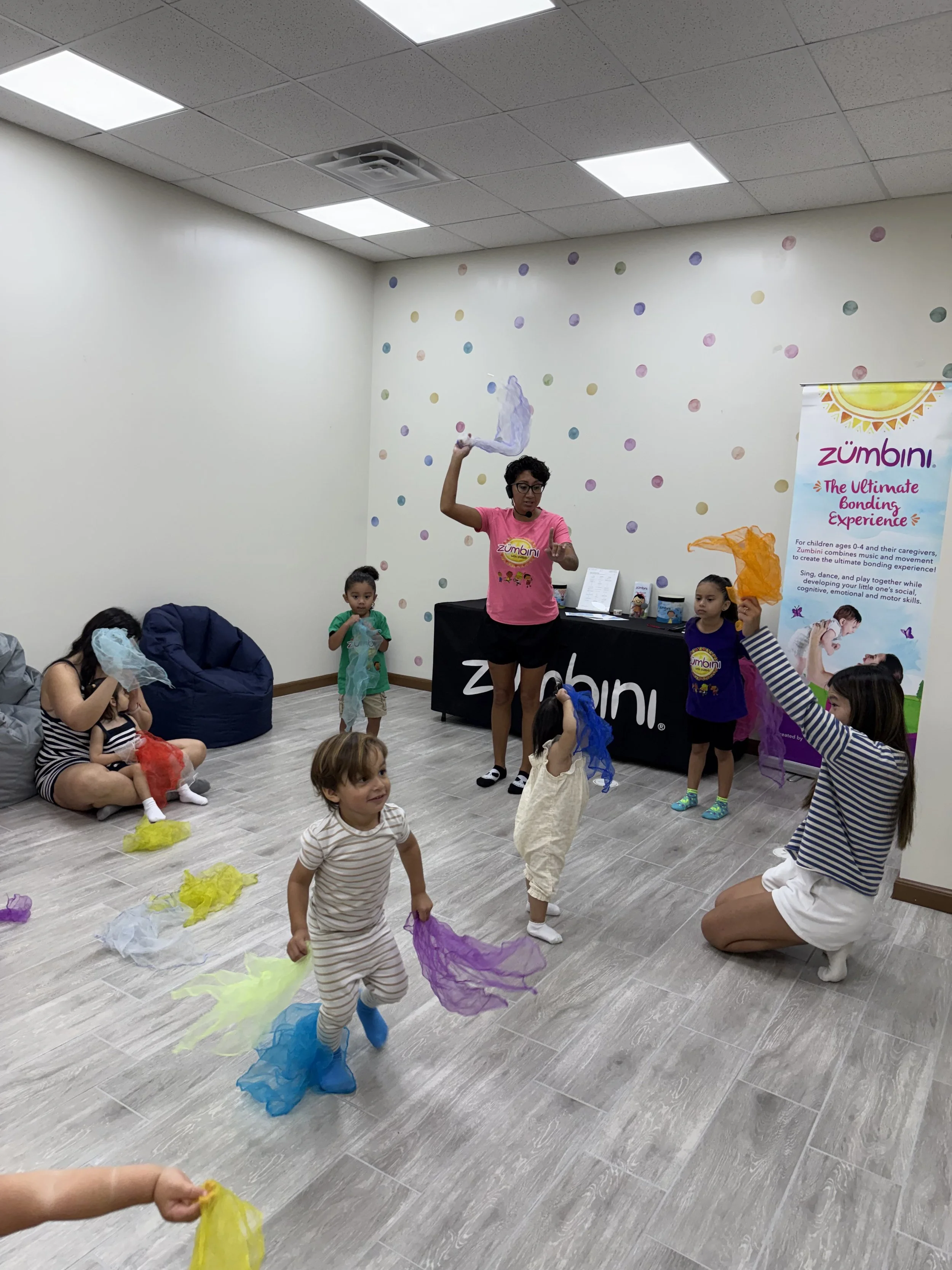 Children and instructors participating in a colorful dance or activity class with scarves in a bright room with a polka dot decorated wall and promotional banner for Zumbini, a music and movement program for young children.