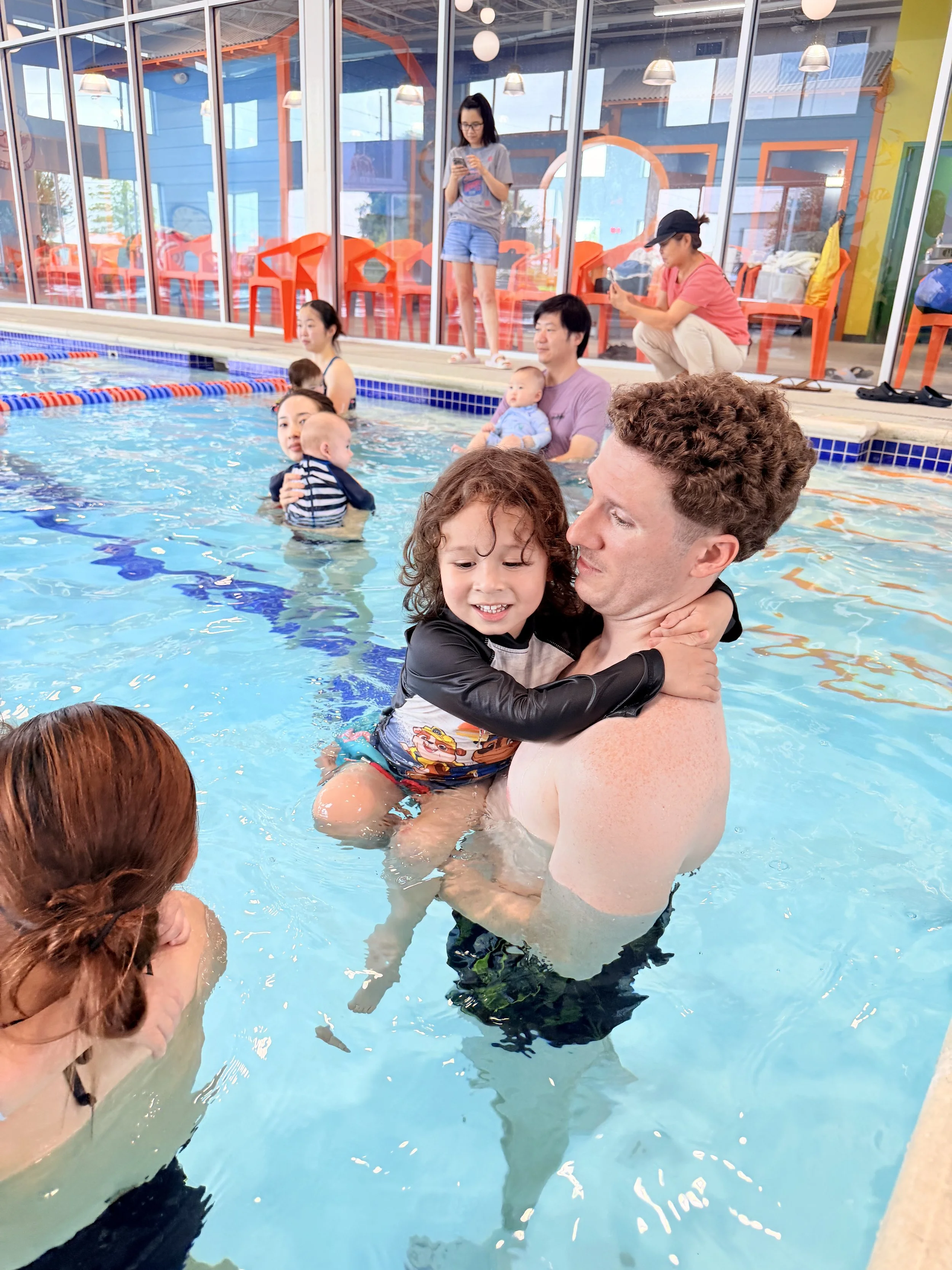 People in a swimming pool, including children and adults, with some standing and holding children, and others sitting or standing by the poolside inside a building with large glass windows and colorful decor.