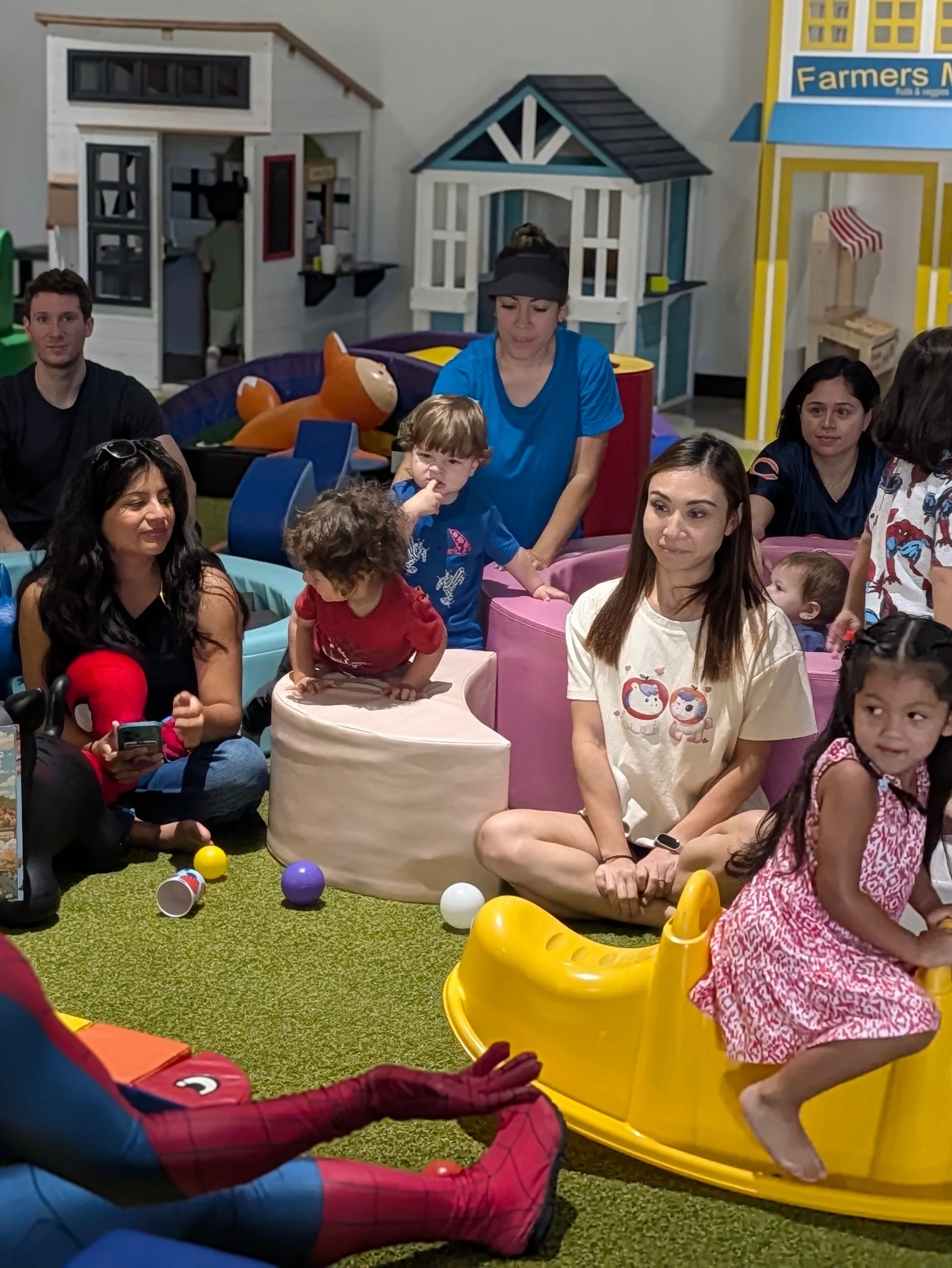 Indoor play area with children and adults, a small house play structure, colorful plastic balls, and a woman with a child at a table.