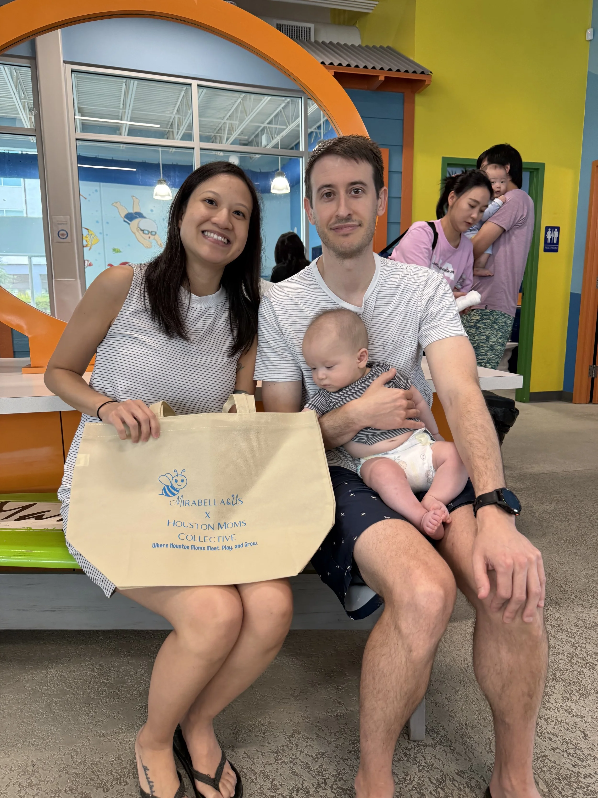 Group of people sitting in a colorful indoor space, including a woman smiling, a man holding a baby, and a woman in the background carrying a child, with a tote bag that has a bee logo and text about Houston moms.