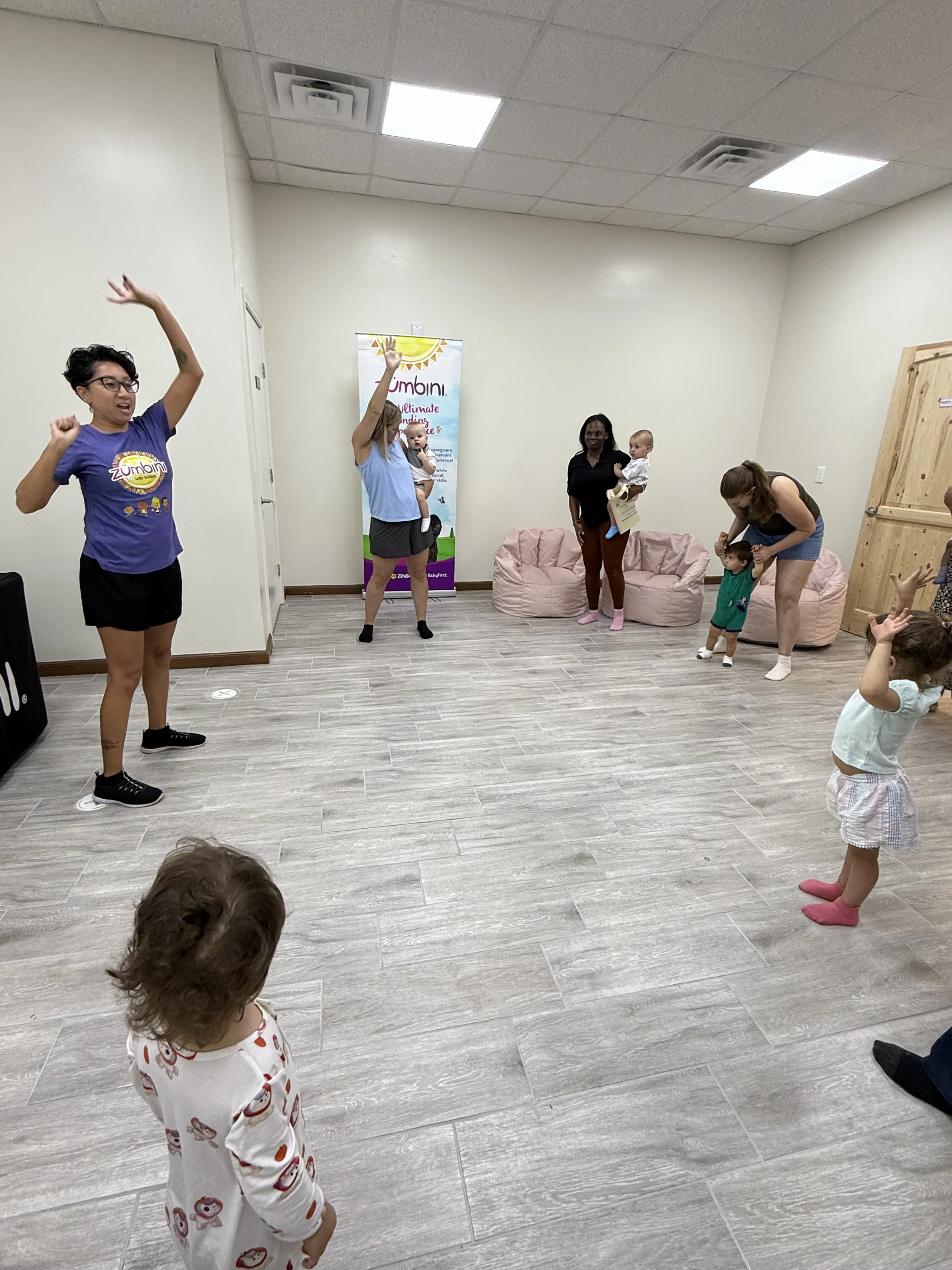 Children and adults participating in a group activity in an indoor room, with soft seating and a banner in the background that reads 'Zumbini: The Ultimate Bonding Experience.'