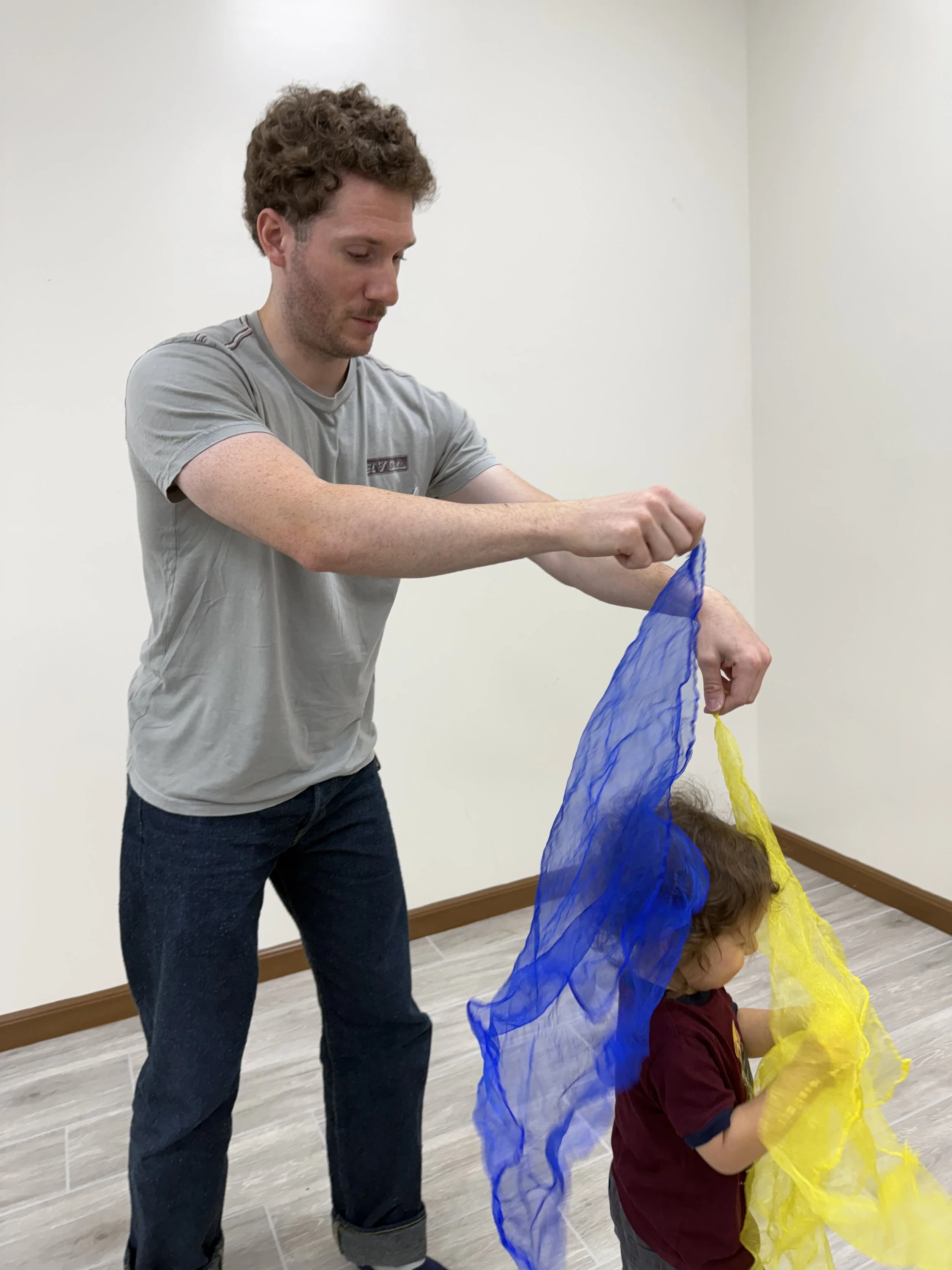 A man helping a child put on a colorful fabric cape in a room with white walls and wood trim.