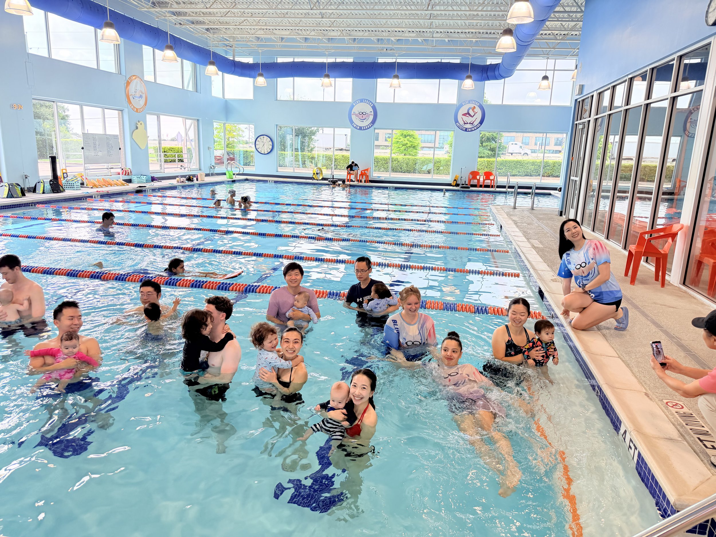 Indoor swimming pool filled with people, including children and adults, enjoying swimming and water activities. There are large windows allowing natural light, with some people sitting and supervising by the edge.