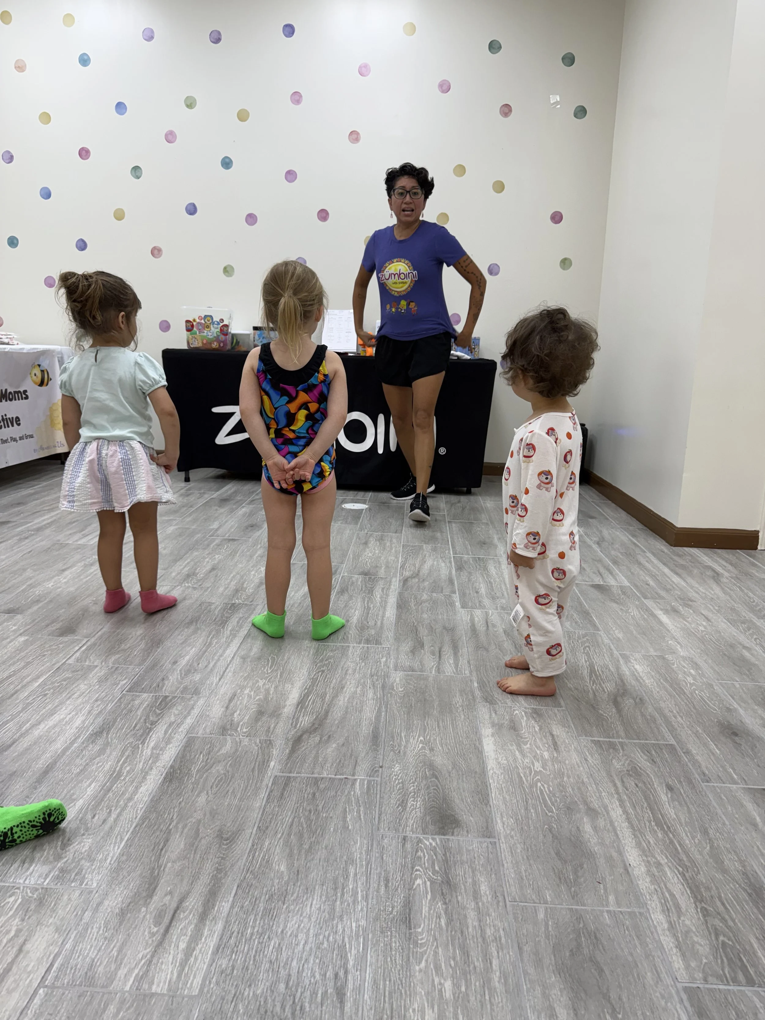 A woman leading a children's exercise class in a room with a polka dot wall, with four children standing in front of her. The woman is wearing glasses, a blue shirt, and black shorts. The children are dressed in colorful pajamas and are barefoot, pay