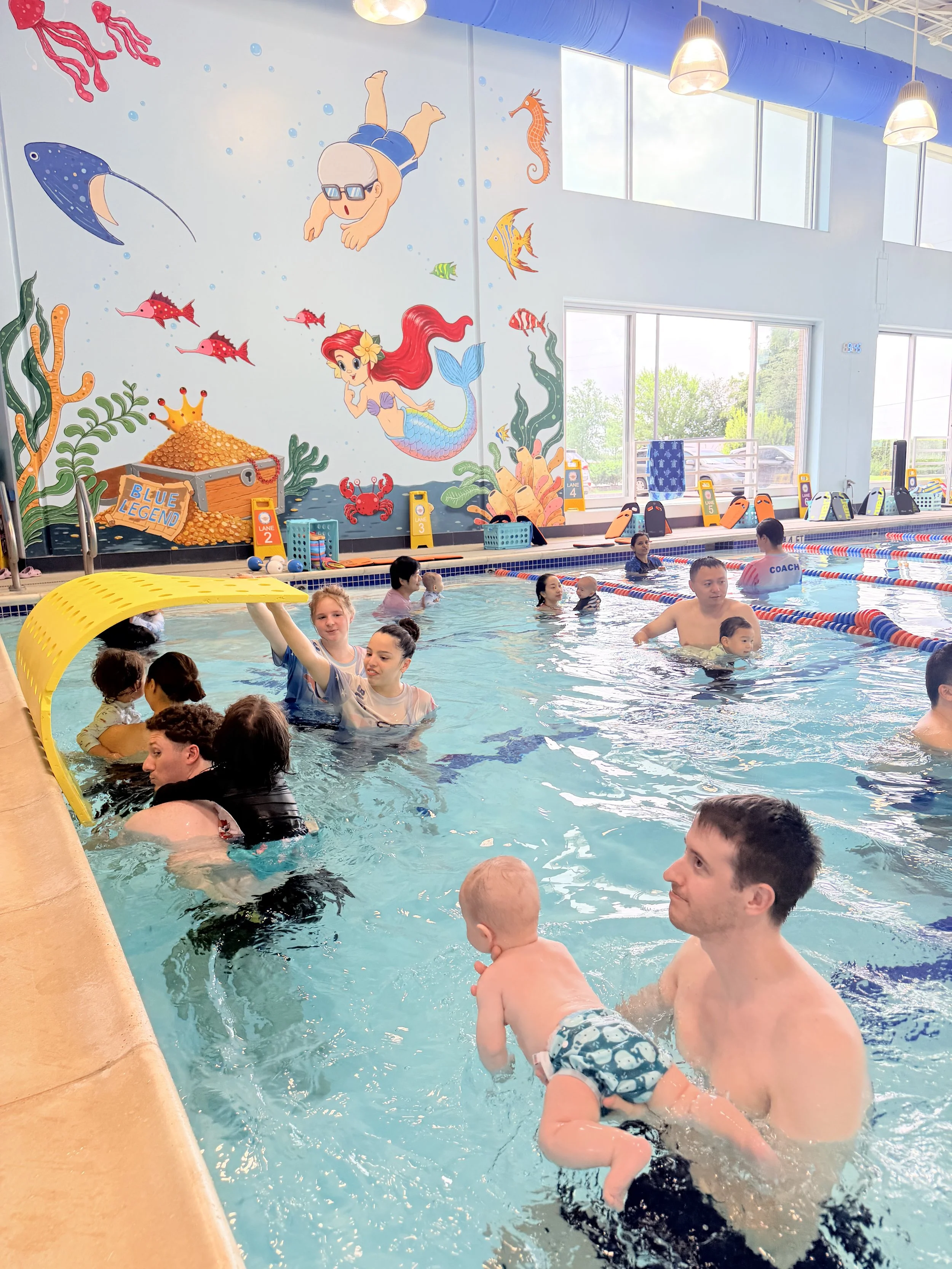 Indoor swimming pool filled with children and adults participating in a swimming lesson, with a colorful underwater-themed mural on the wall featuring a mermaid, a man swimming, various fish, a crab, a seahorse, seaweed, and a treasure chest.