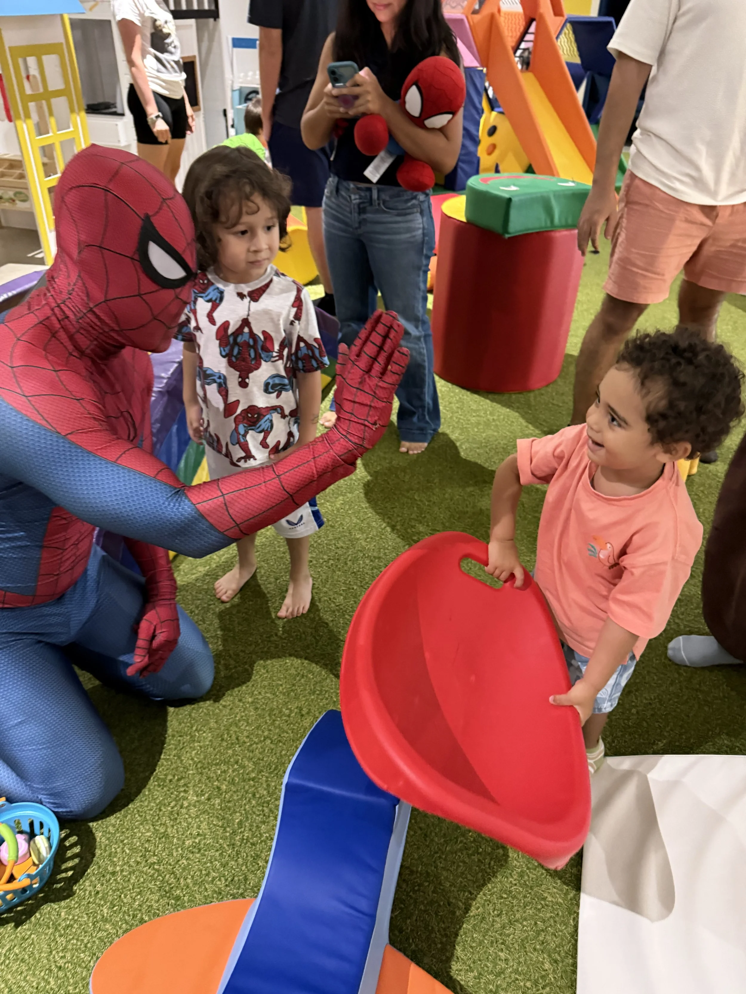 A person dressed as Spider-Man kneels in front of two young children in a play area, possibly at a toy or game store, with one of the children holding a red plastic disc.