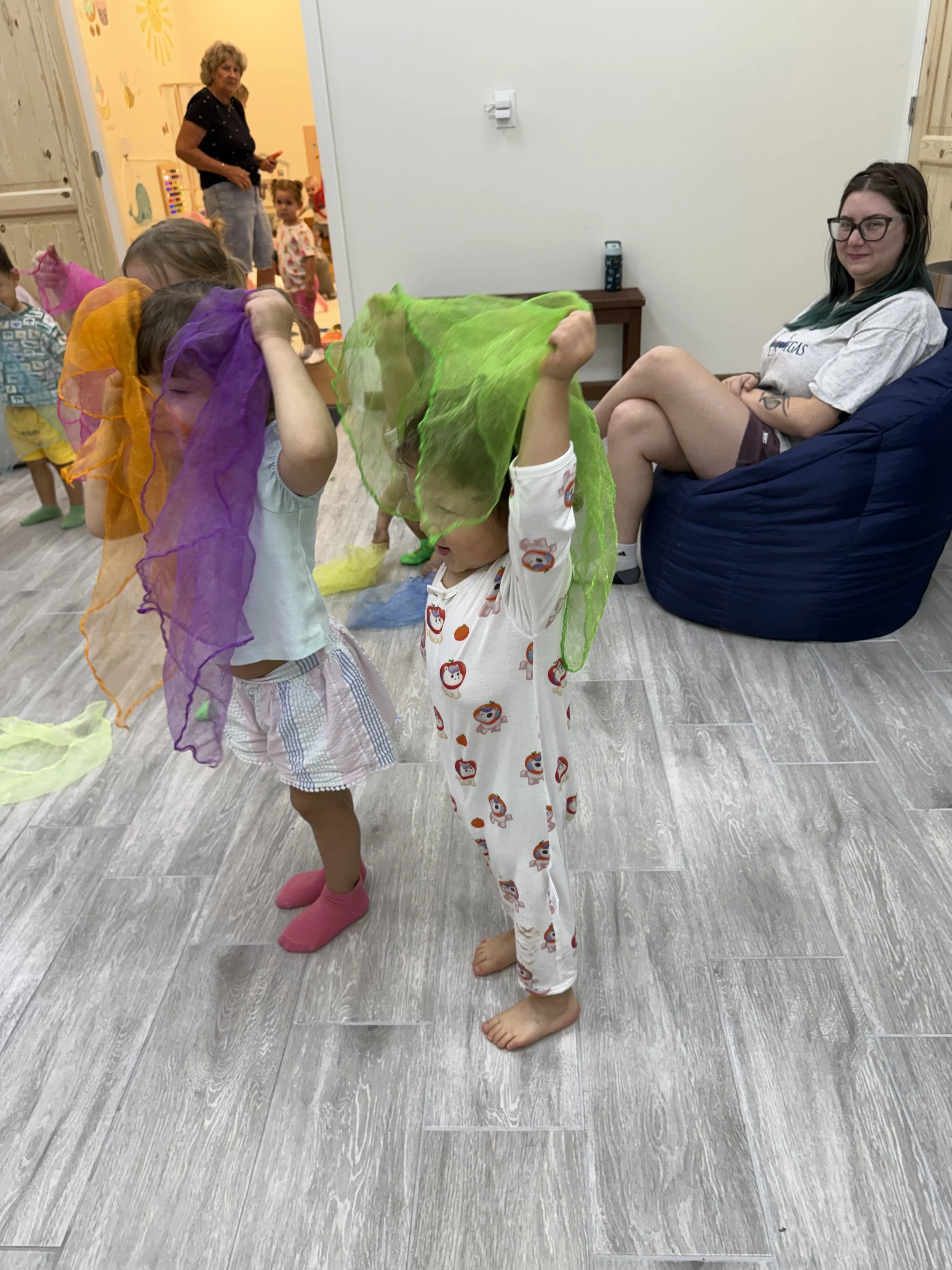 Children playing with colorful scarves in a room, a woman sitting on a bean bag, and a woman standing in the background in a room decorated with playful wall art.