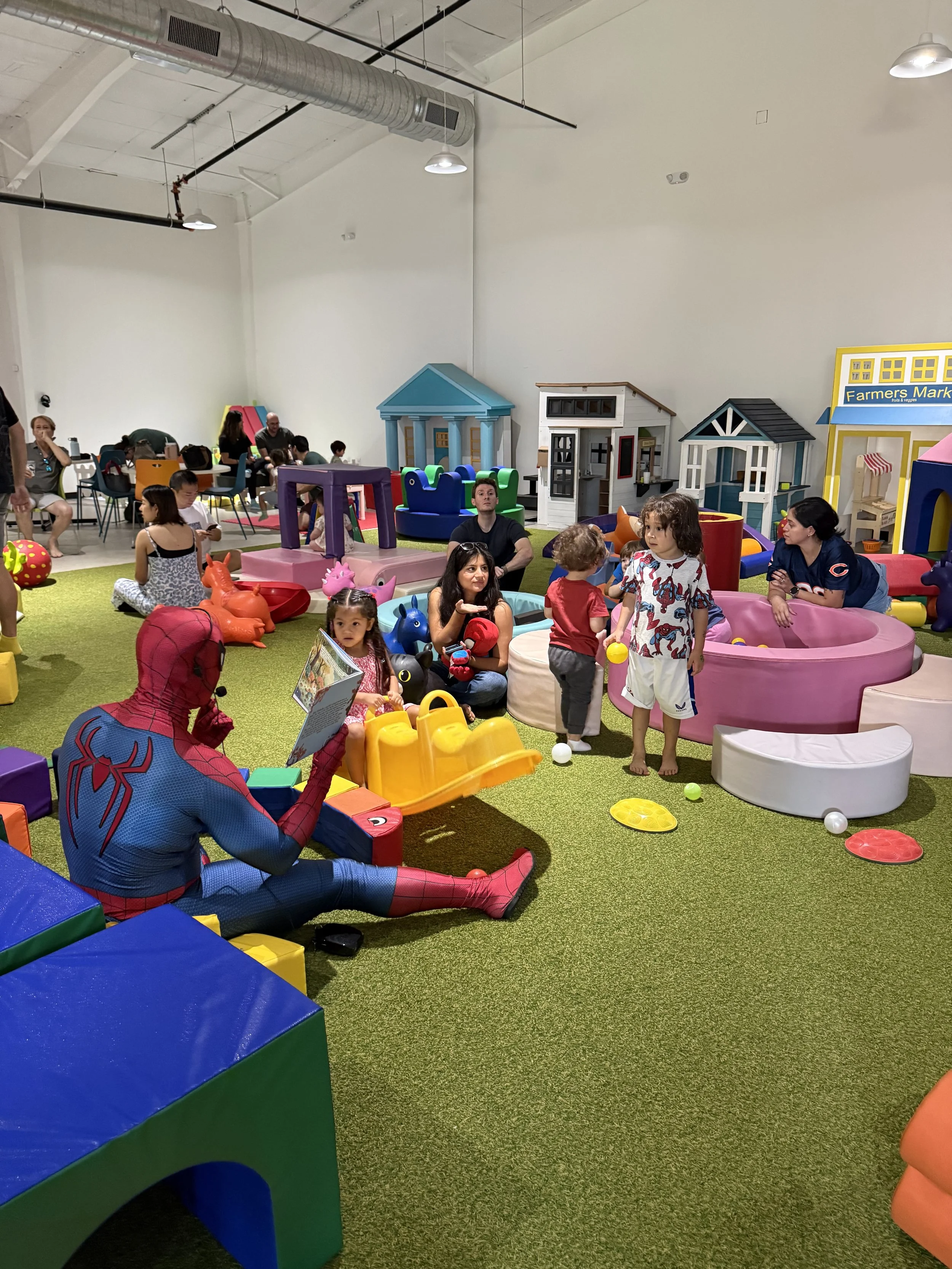 Indoor children's play area with various colorful soft play structures, toys, and some adults and children playing and interacting. A person dressed as Spider-Man is sitting on the floor reading a book.