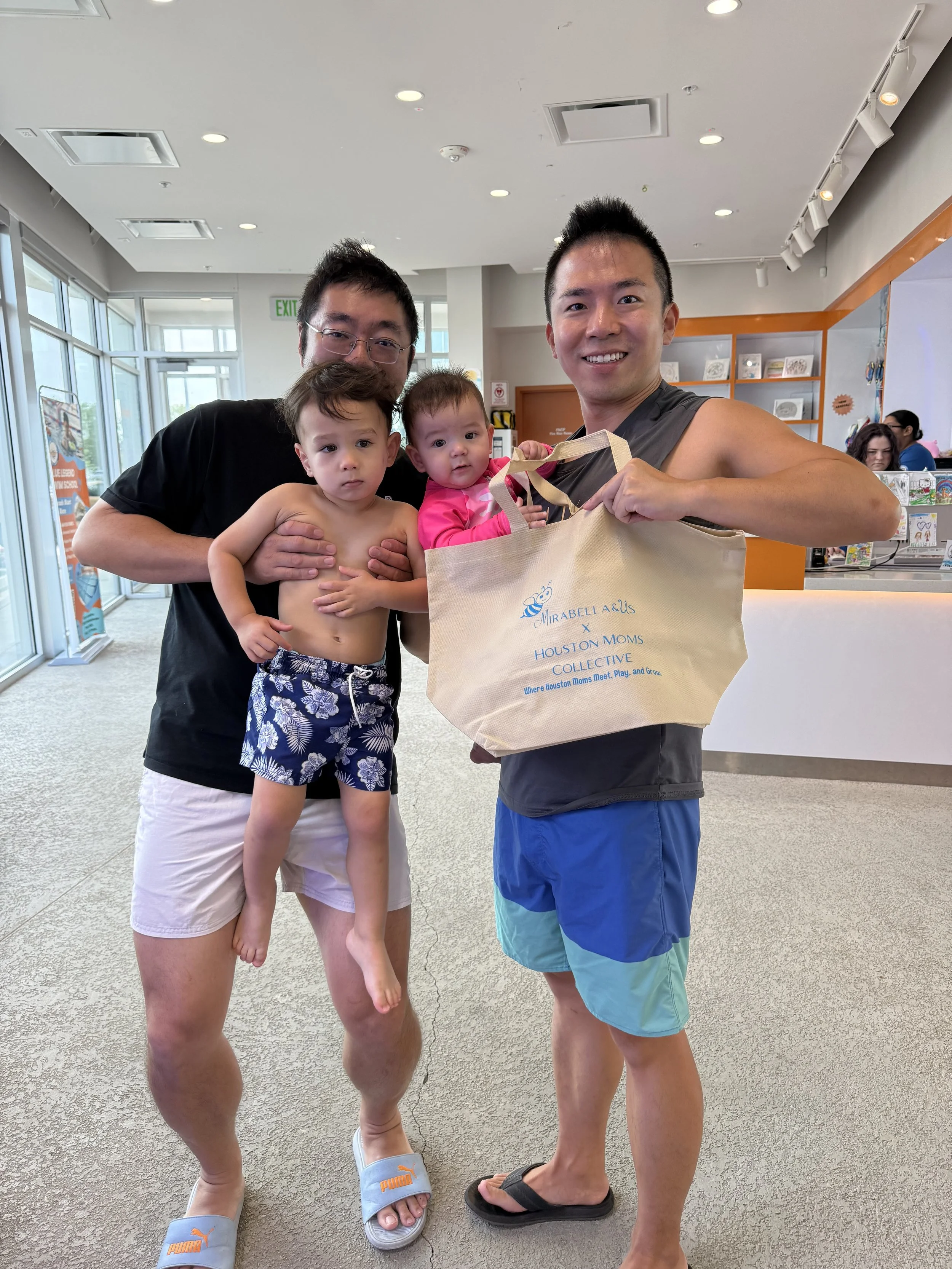 Two men and two young children smiling inside a brightly lit indoor space with large windows, shelves with books and artwork, and a front desk area.