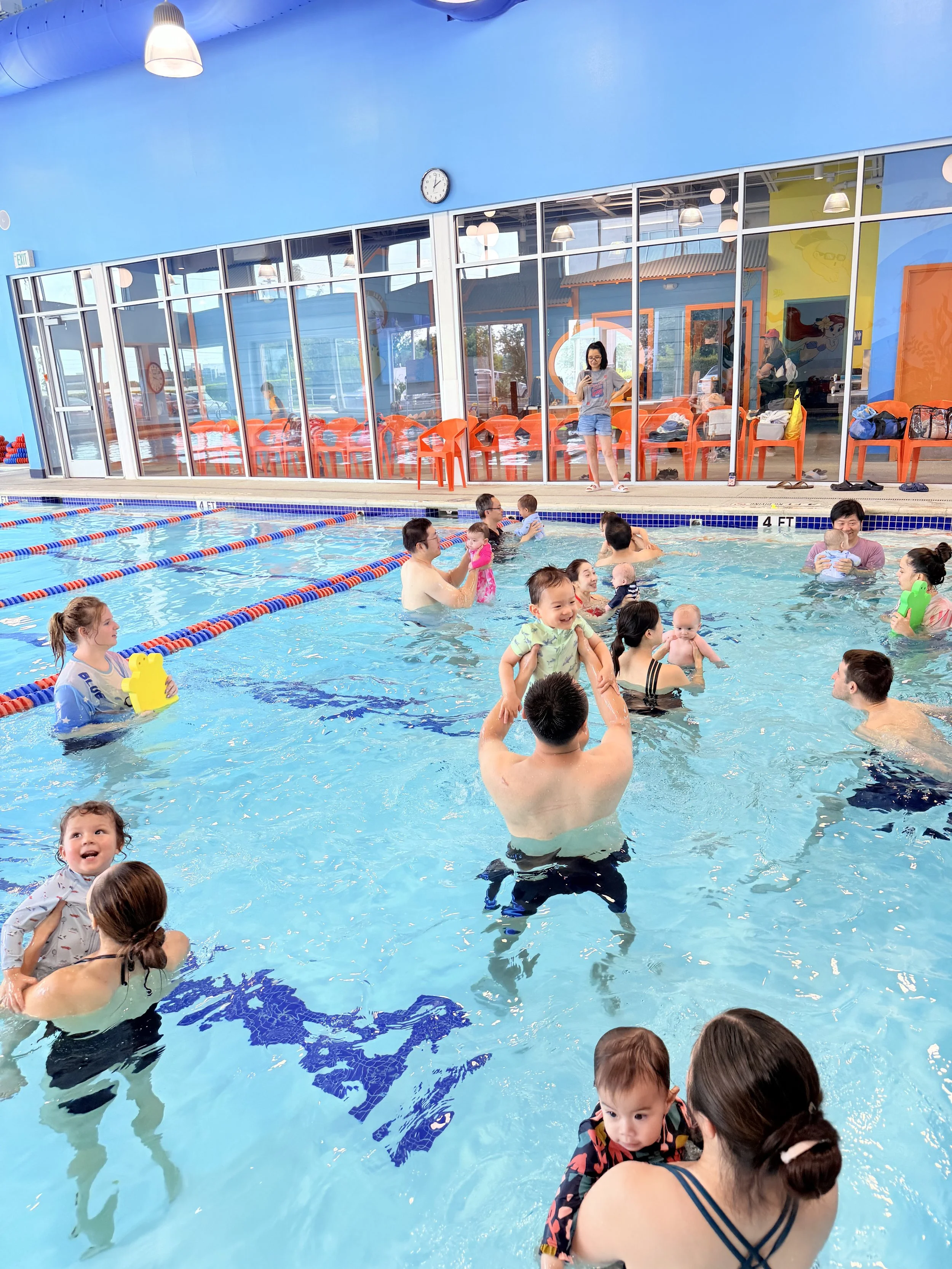 Indoor swimming pool filled with children and adults enjoying swimming and water activities, with colorful walls and glass windows in the background.