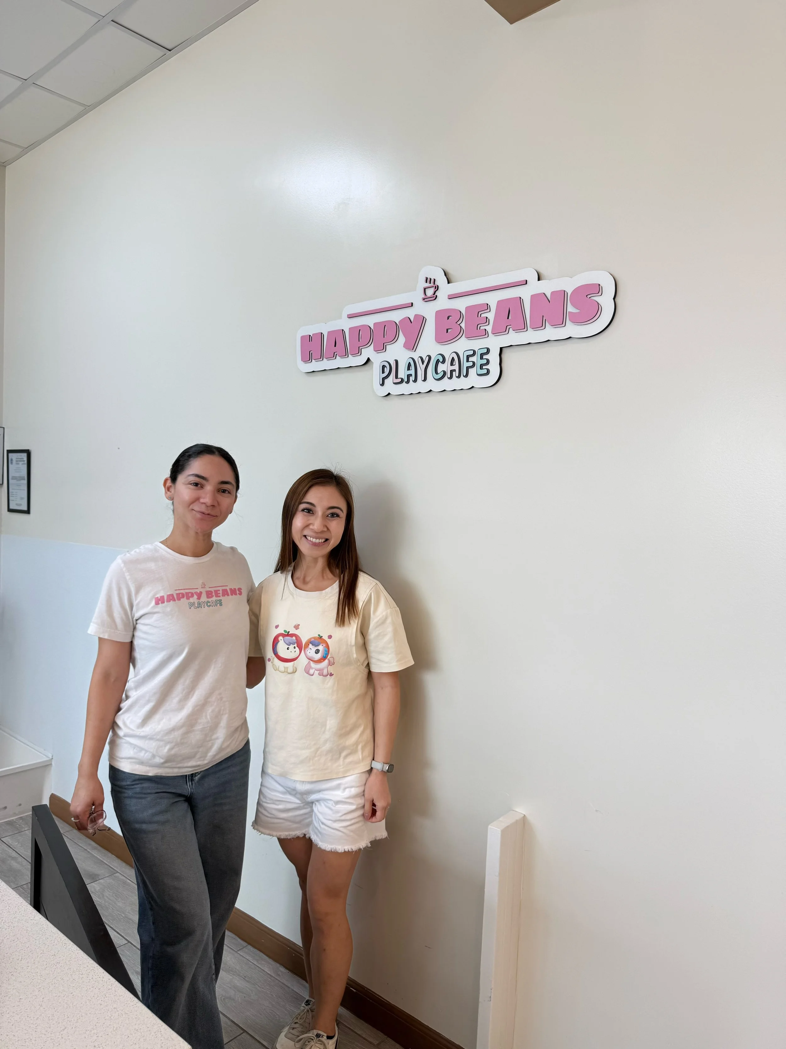 Two women standing next to each other indoors, smiling, in front of a wall with a sign that reads 'Happy Beans Play Cafe'.