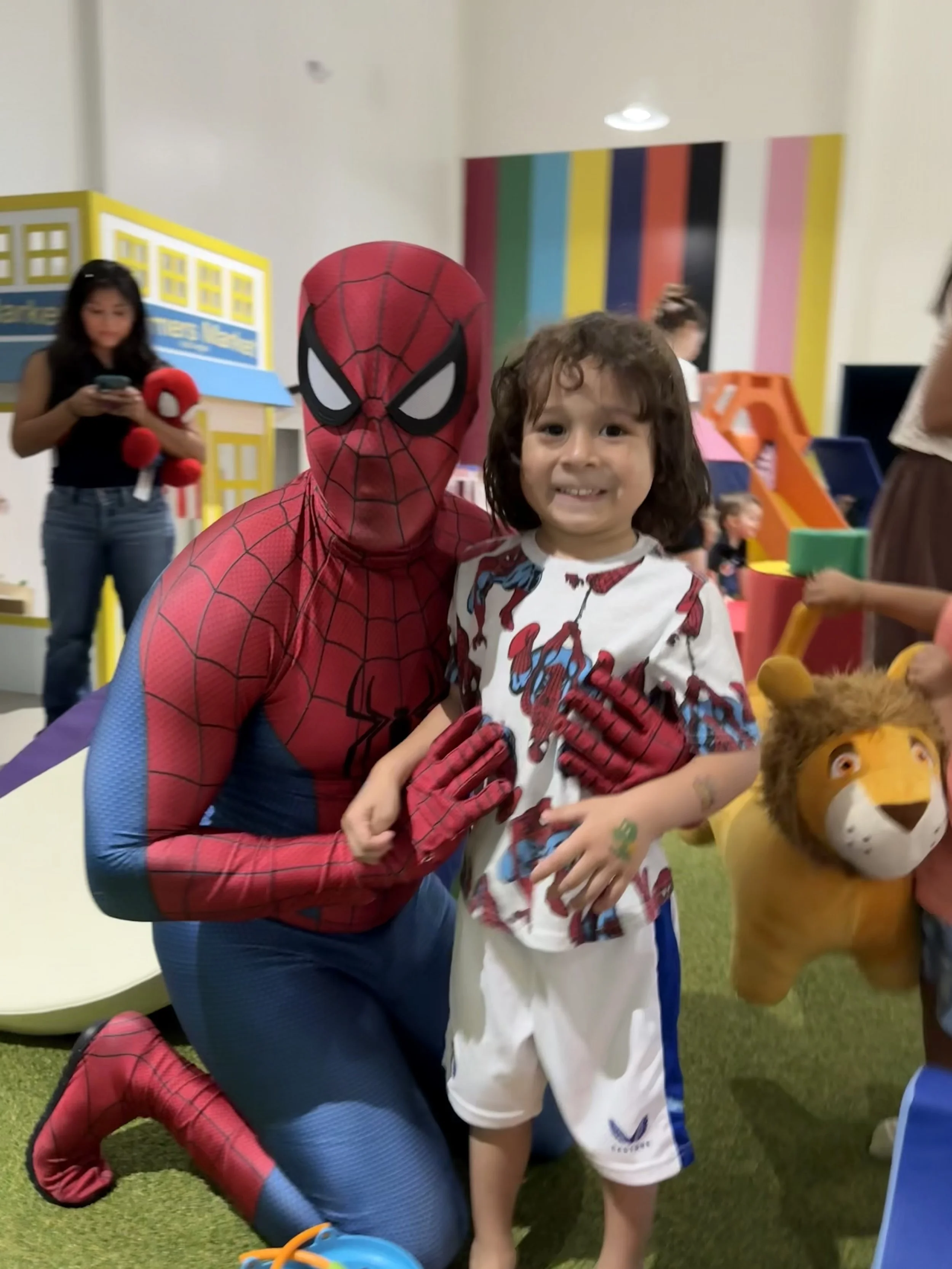 A person dressed as Spider-Man kneeling next to a young boy smiling at the camera, in a play area with colorful walls and toys.