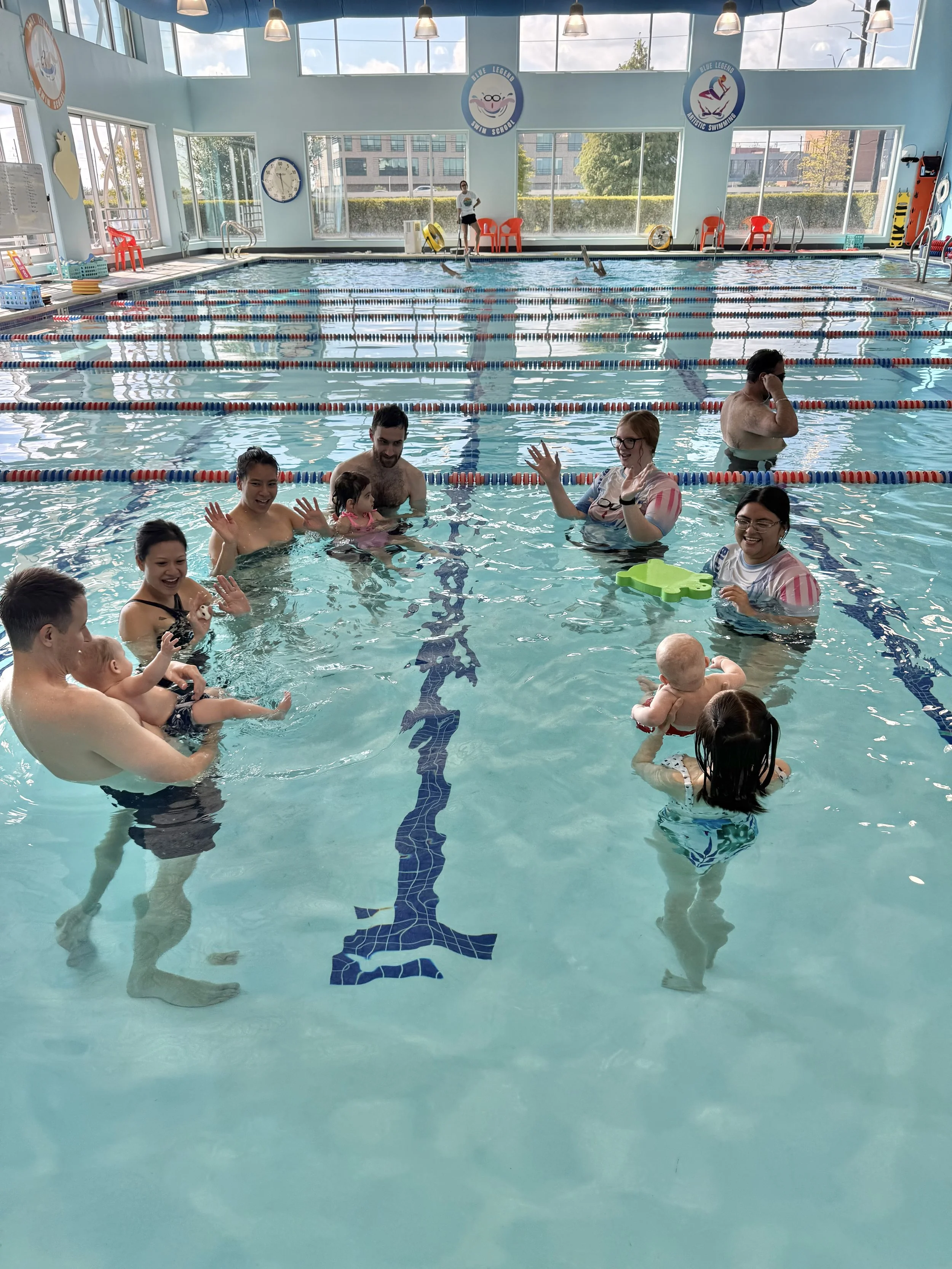 Indoor swimming pool with children and adults enjoying swimming lessons. Bright room with large windows and blue themed decorations.