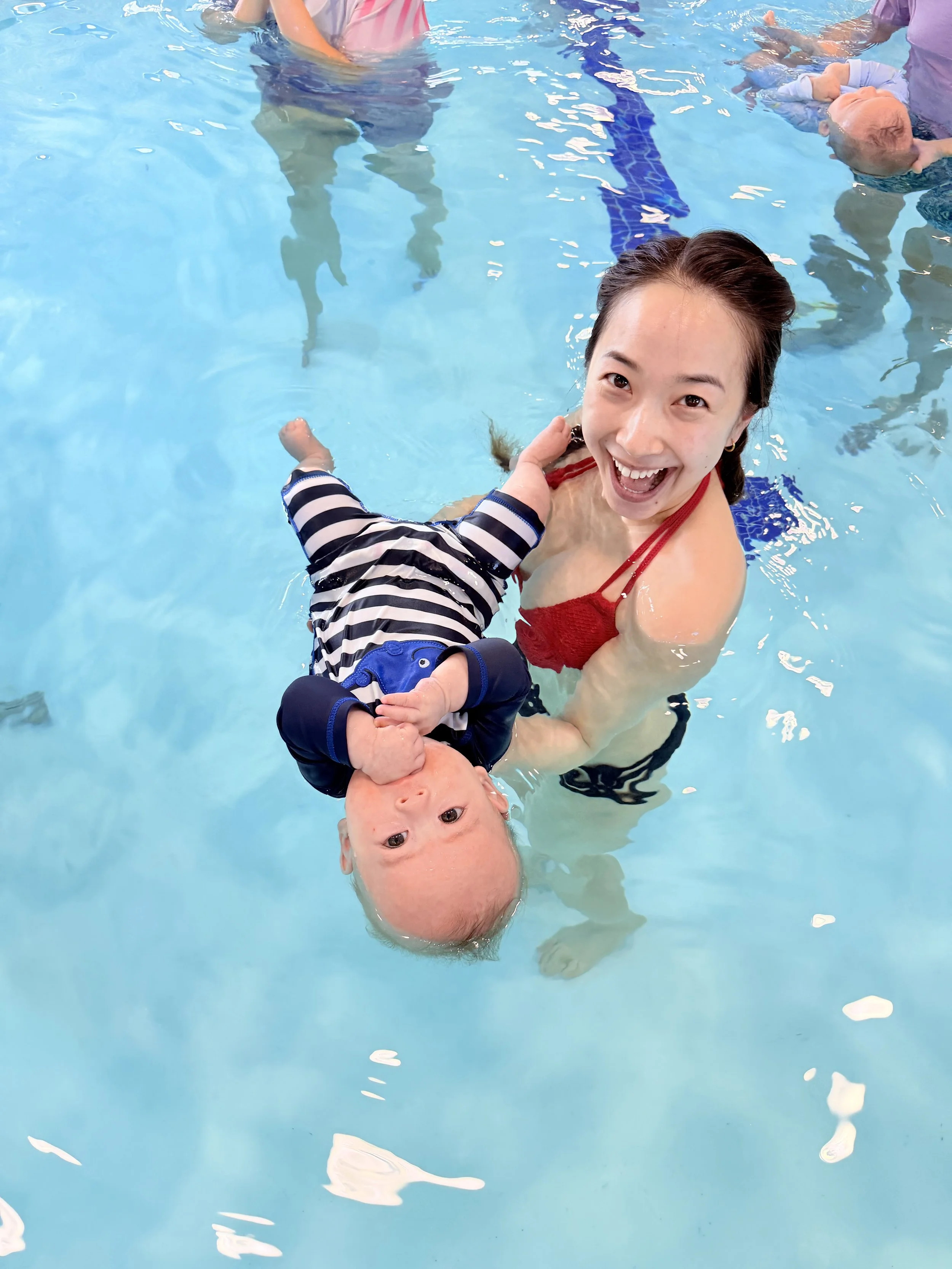 A woman in a red swimsuit holding a bald, smiling baby in striped clothing in a swimming pool with other children and adults in the background.