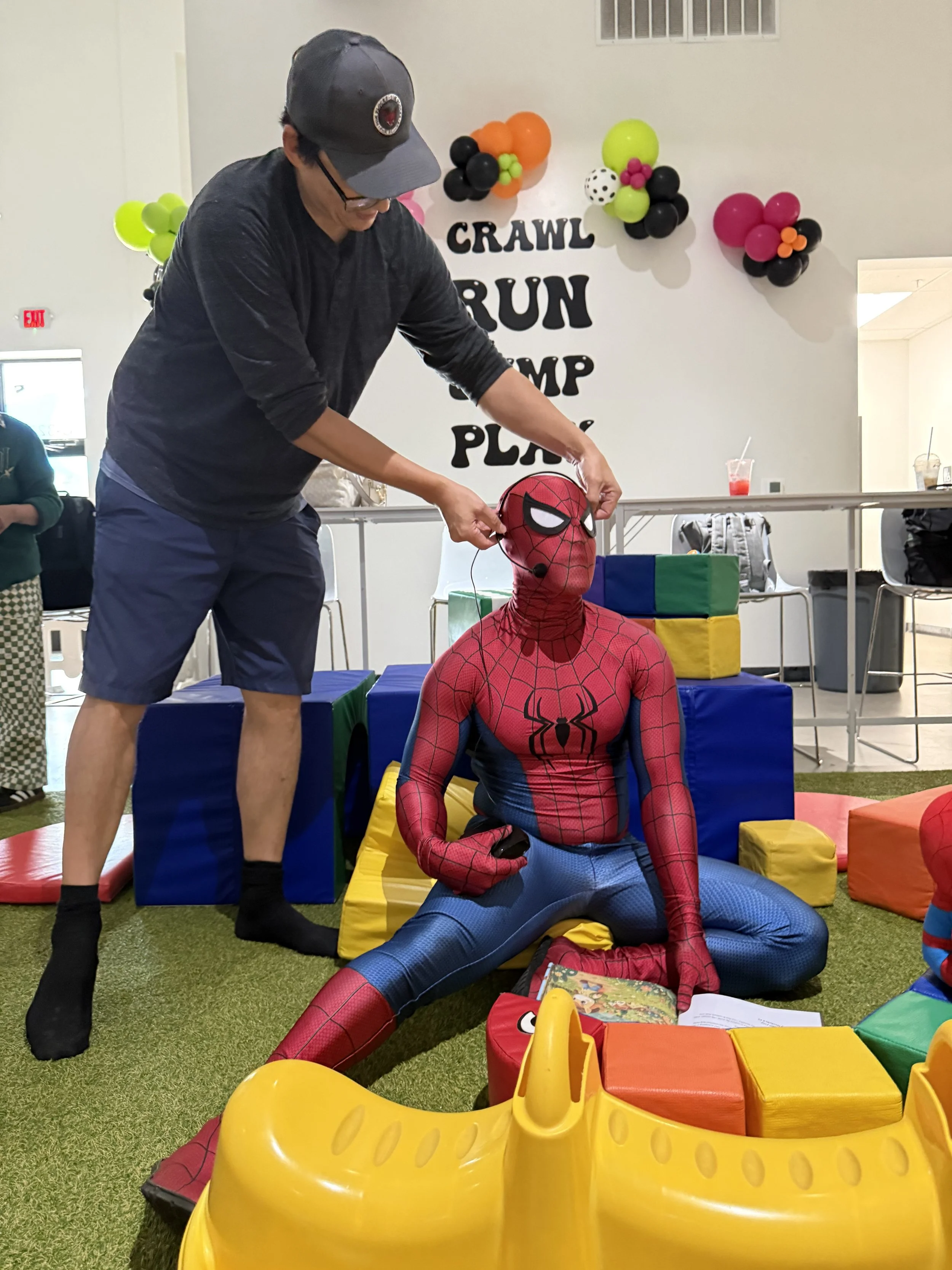 A person dressed as Spider-Man sitting on the floor amid colorful foam mats and soft play blocks, with an adult adjusting a Spider-Man mask on a head mannequin at a children's play area decorated with balloons and a sign reading 'Crawl, Run, Jump, Pl