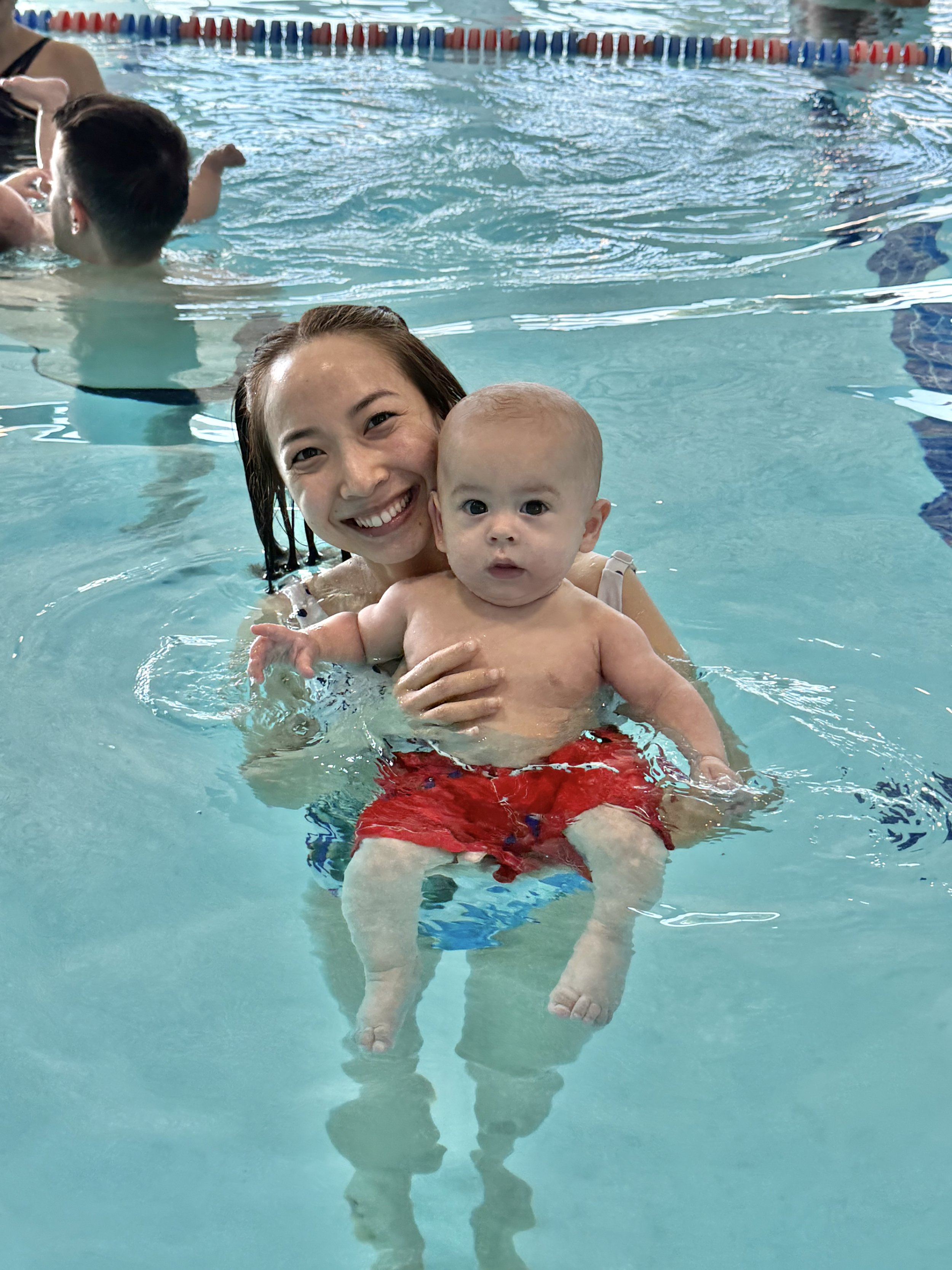 A woman smiling while holding a young child in a swimming pool, with other children swimming nearby.