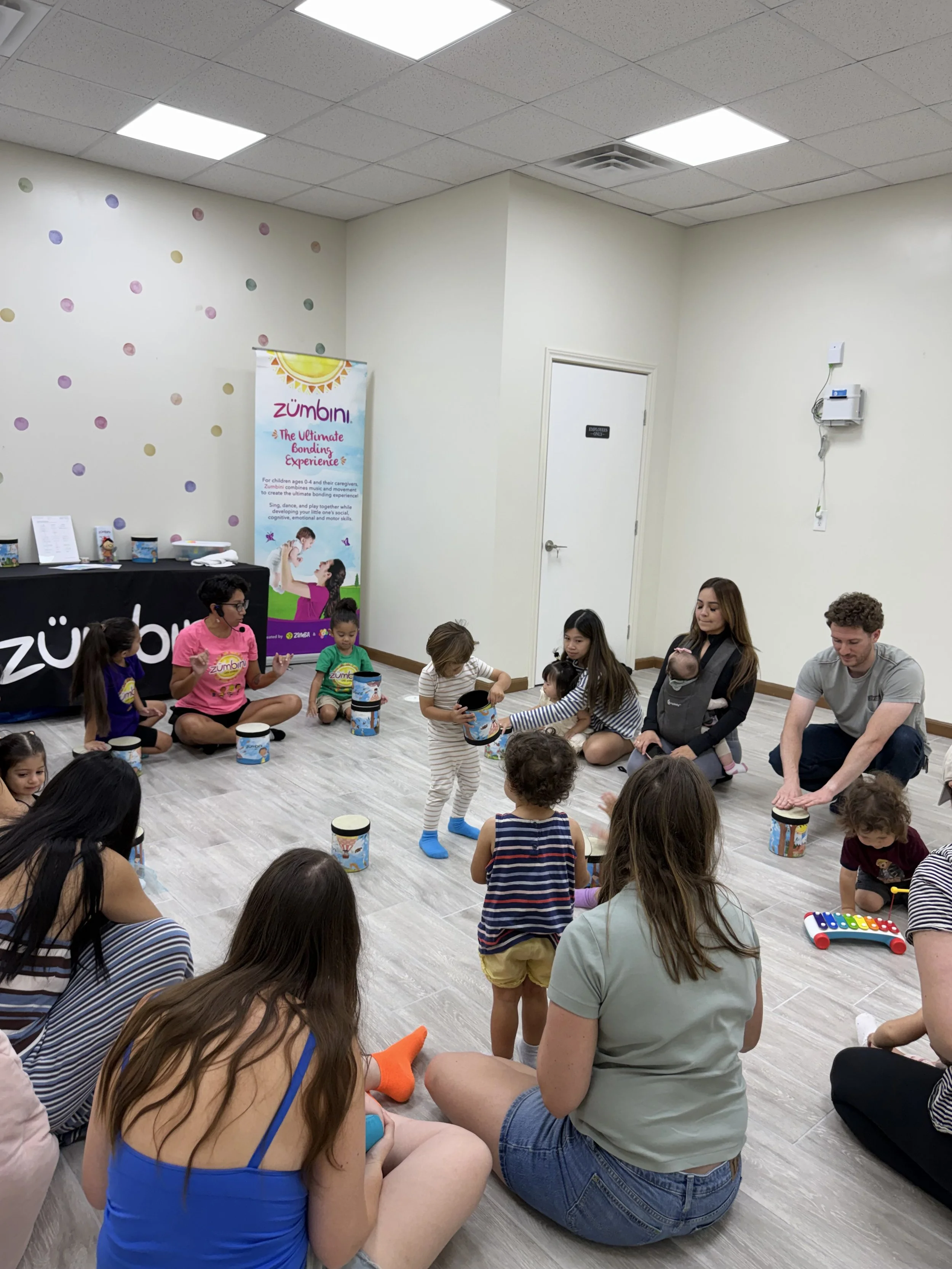 Children and adults participating in a children's activity class, sitting on the floor with drums and musical toys, in a brightly lit room with plain walls and ceiling tiles.