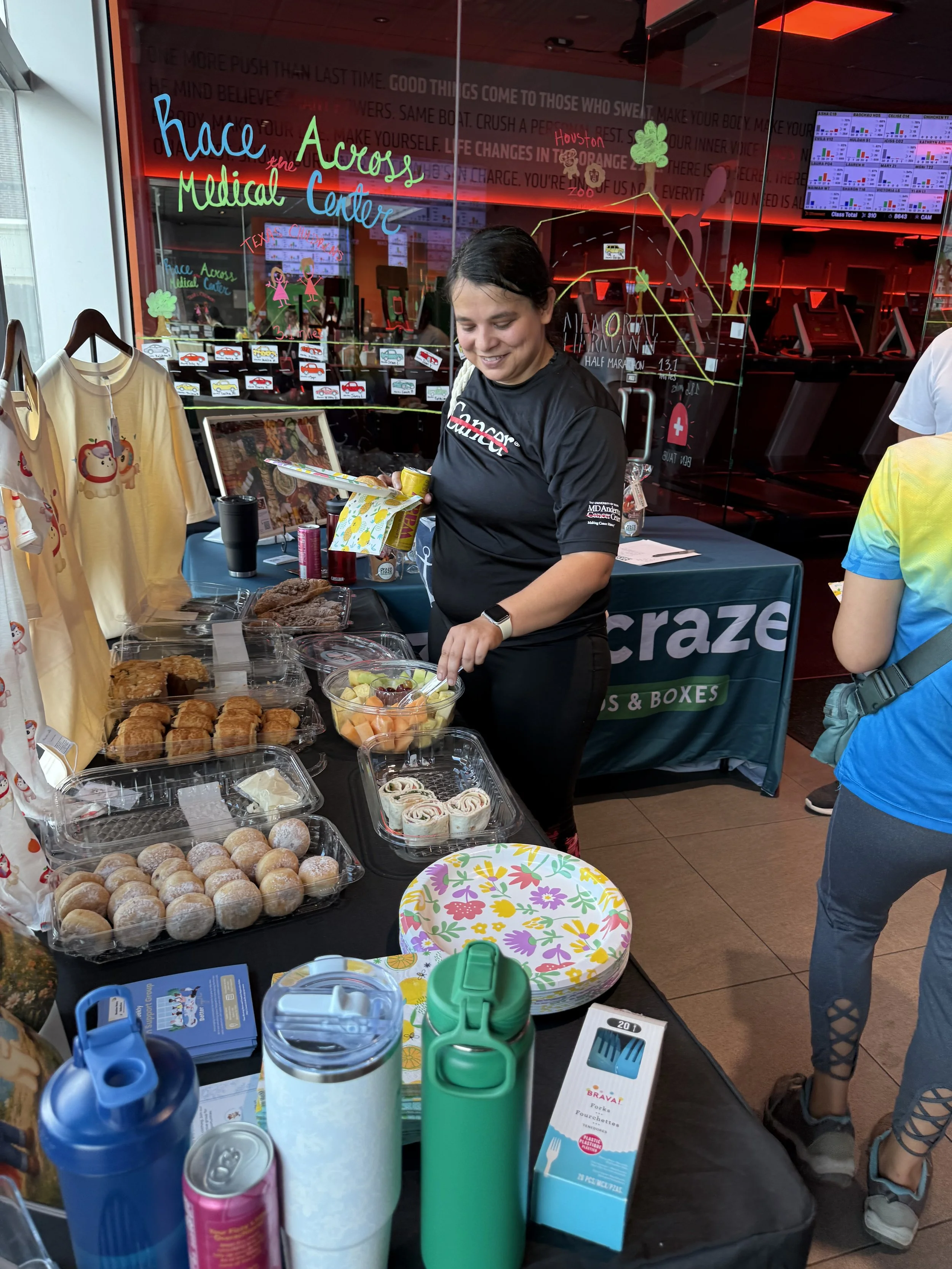 A woman smiling and serving herself fruit salad at a bake sale table with various baked goods, water bottles, and paper plates. Behind her, there is a glass wall with colorful writing and decorations, and a reflection of a treadmill area.