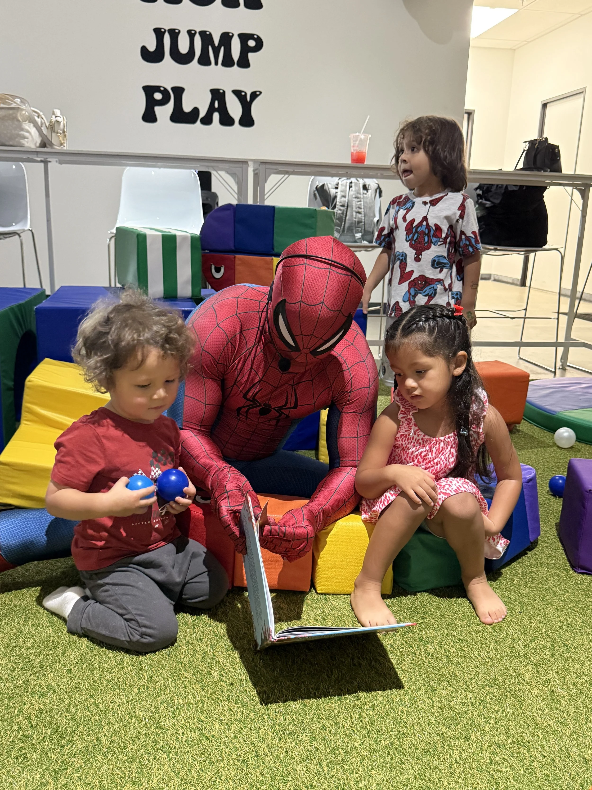 Children sitting on colorful foam mats and a person dressed as Spider-Man at a play area with a sign that says "JUMP PLAY" in the background.