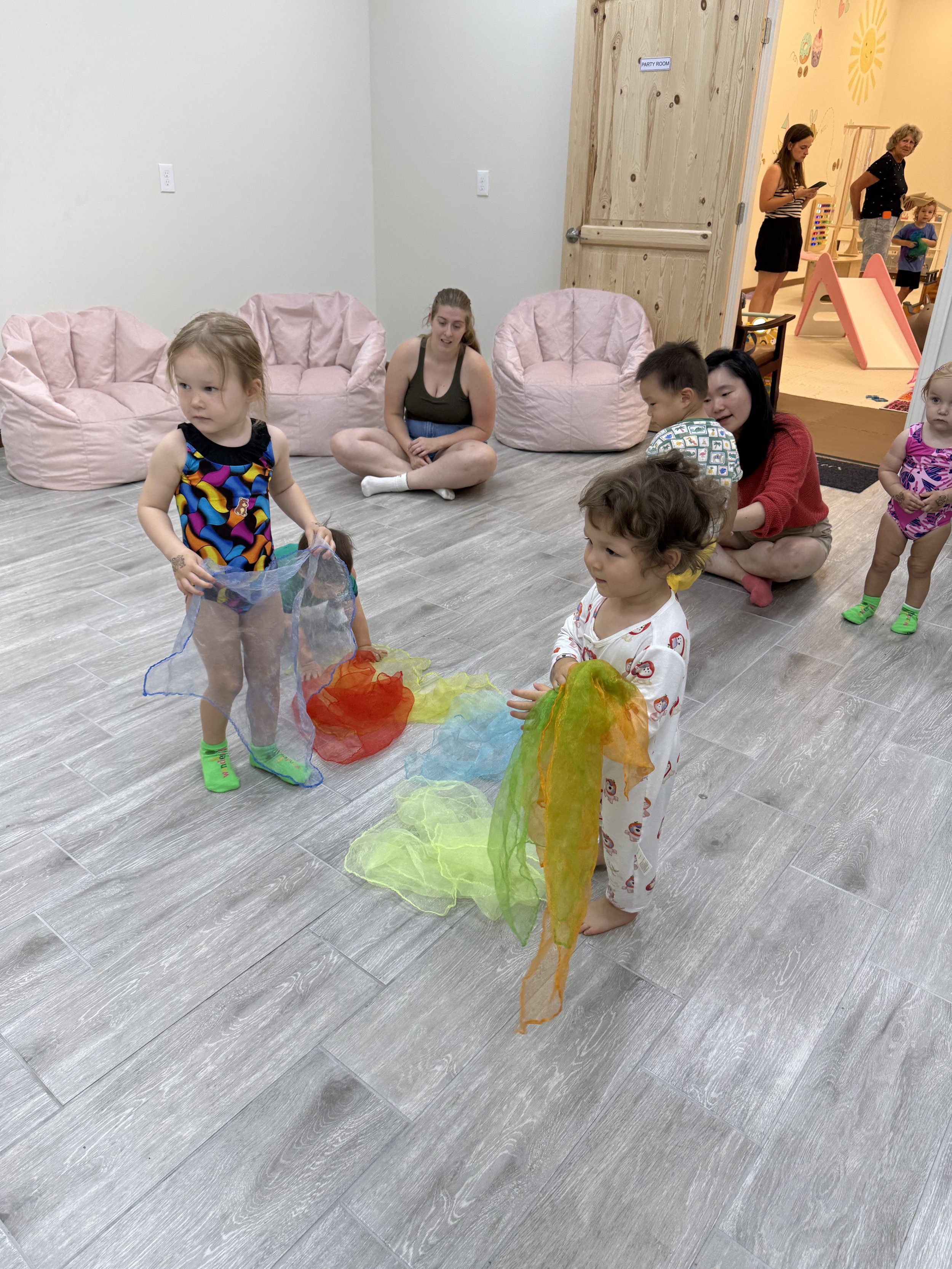 Children playing with colorful fabric scarves on the floor of a playroom as adults watch and supervise, with pink cushioned chairs against the wall and an open door leading to a room with toys and a slide.