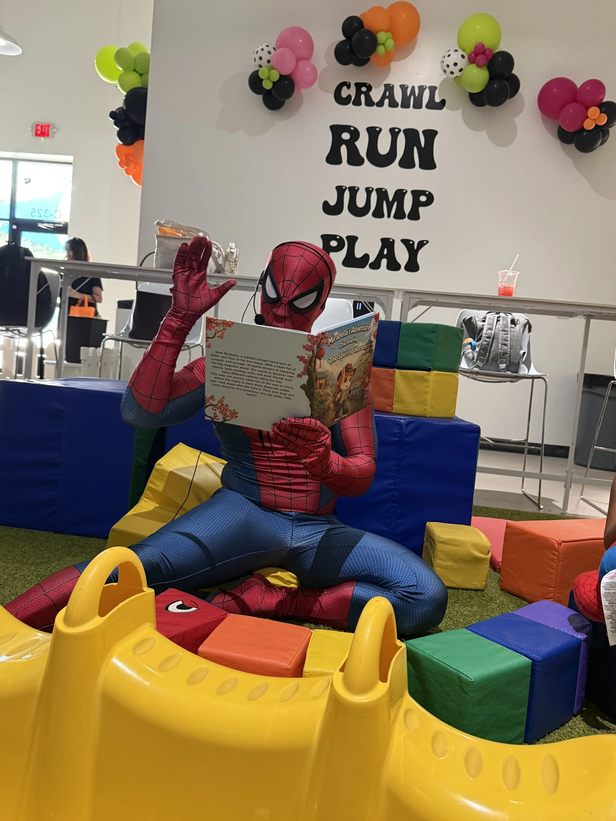 Person dressed as Spider-Man sitting on colorful foam blocks in a play area with balloons and signs reading "Crawl, Run, Jump, Play."