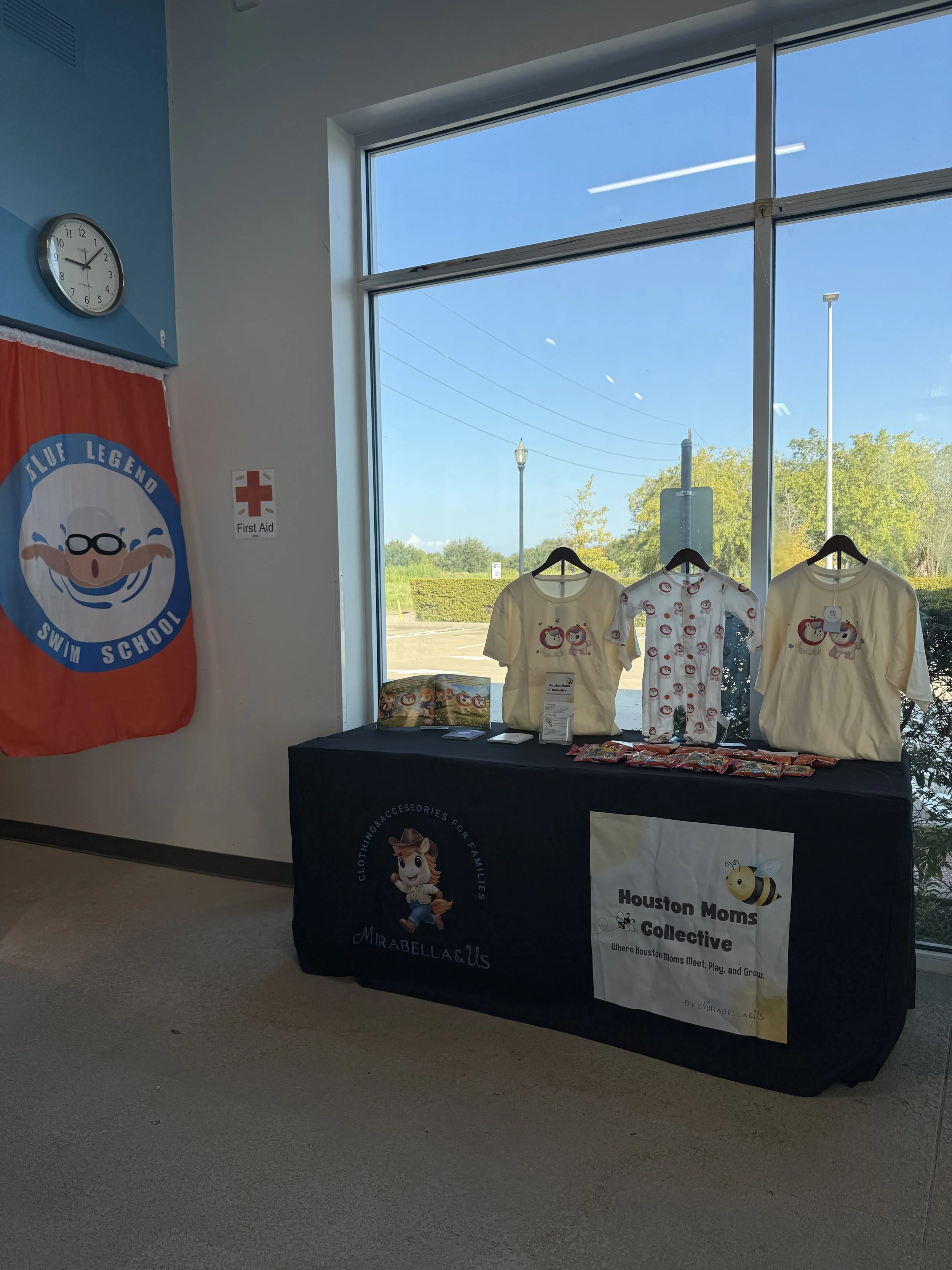 Indoor display table for Houston Moms Collective featuring children's t-shirts with cartoon animal designs, brochures, and a sign with the group's logo, next to a large window with a view of the outdoors.
