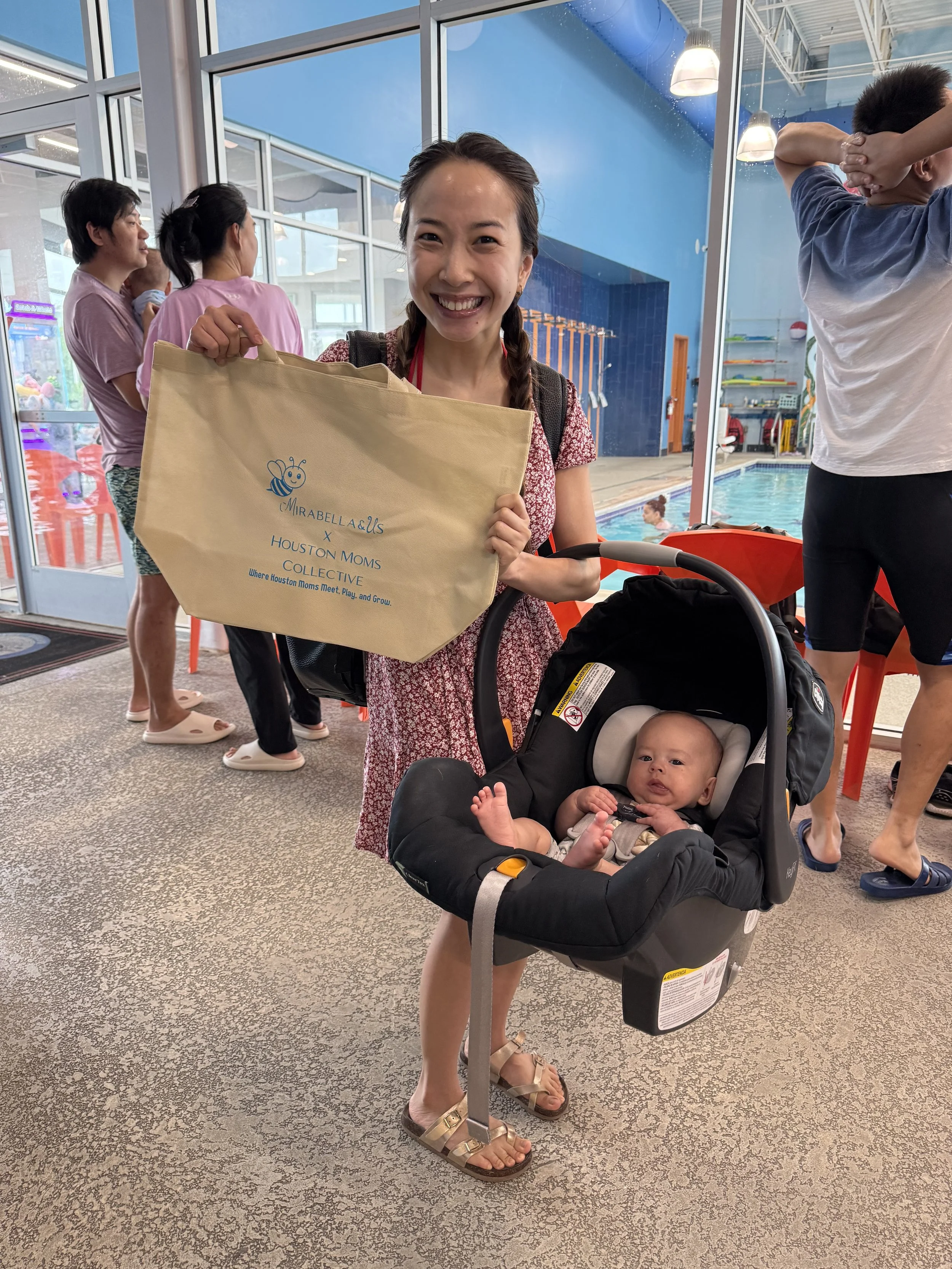 A woman smiling and holding a tote bag with a baby in a car seat stroller at an indoor aquatic center. Other people are in the background near a swimming pool.