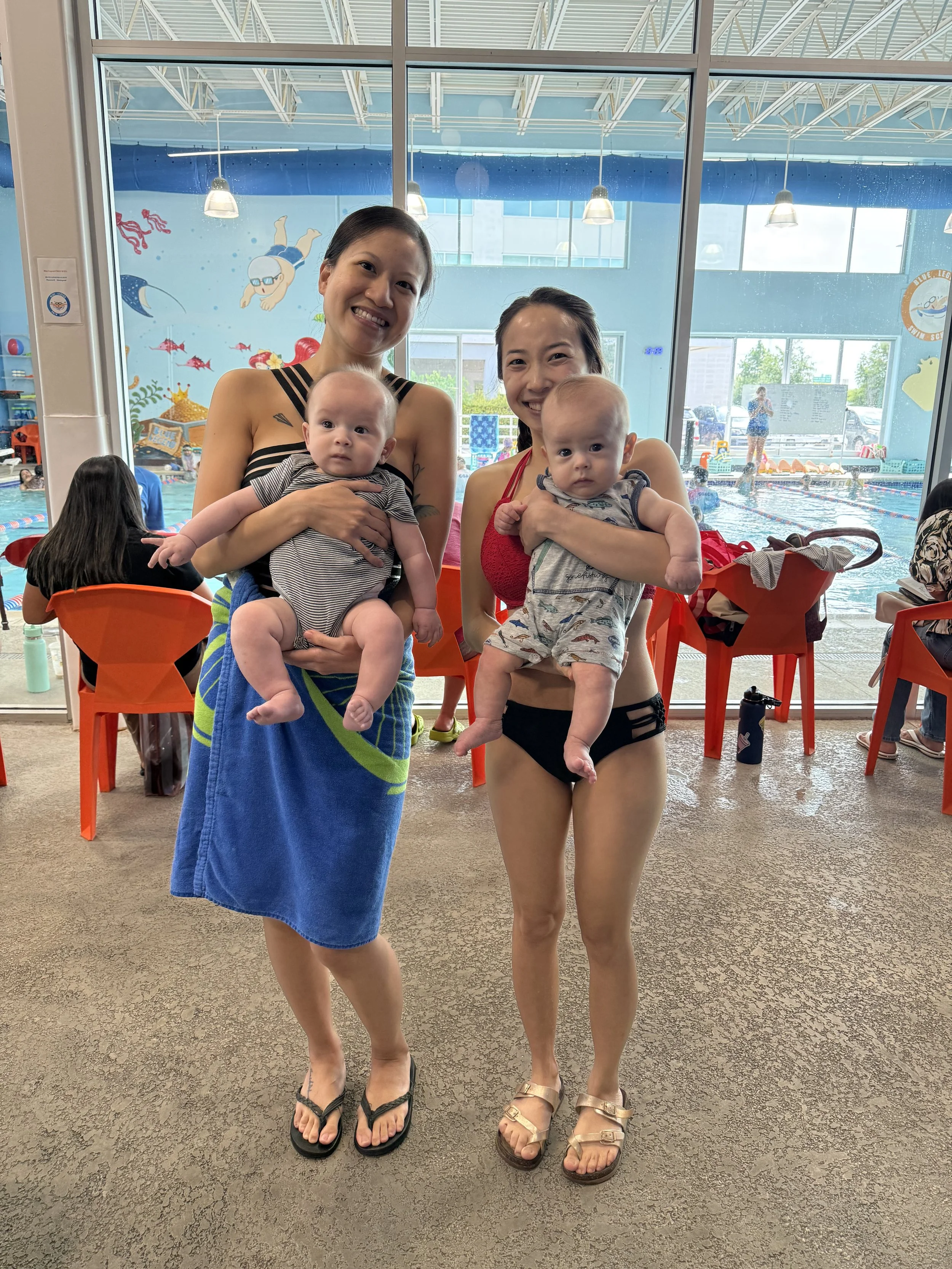 Two women standing in front of a large window at an indoor swimming pool, each holding a baby. The woman on the left has black hair, is smiling, and is wrapped in a blue towel. The woman on the right has dark hair, is smiling, and is wearing a red sw